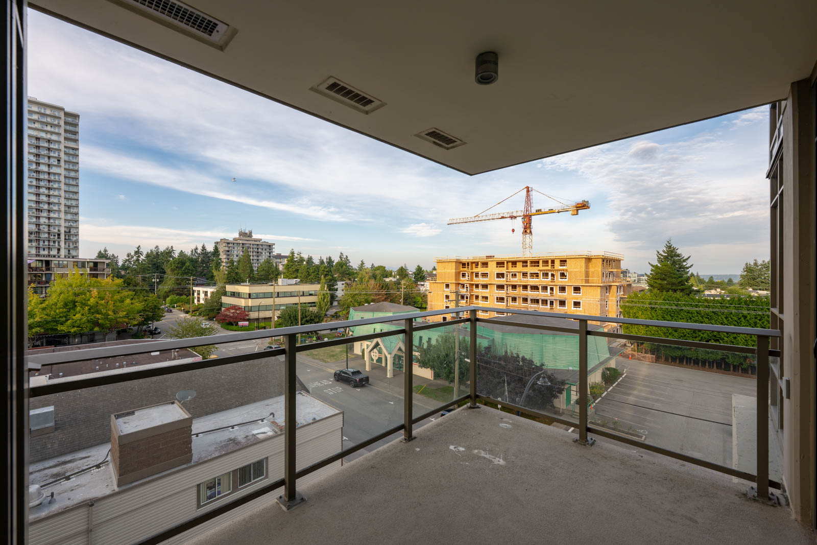 View from a covered balcony with glass railing overlooking a construction site, trees, a parking lot, and several buildings under a partly cloudy sky.