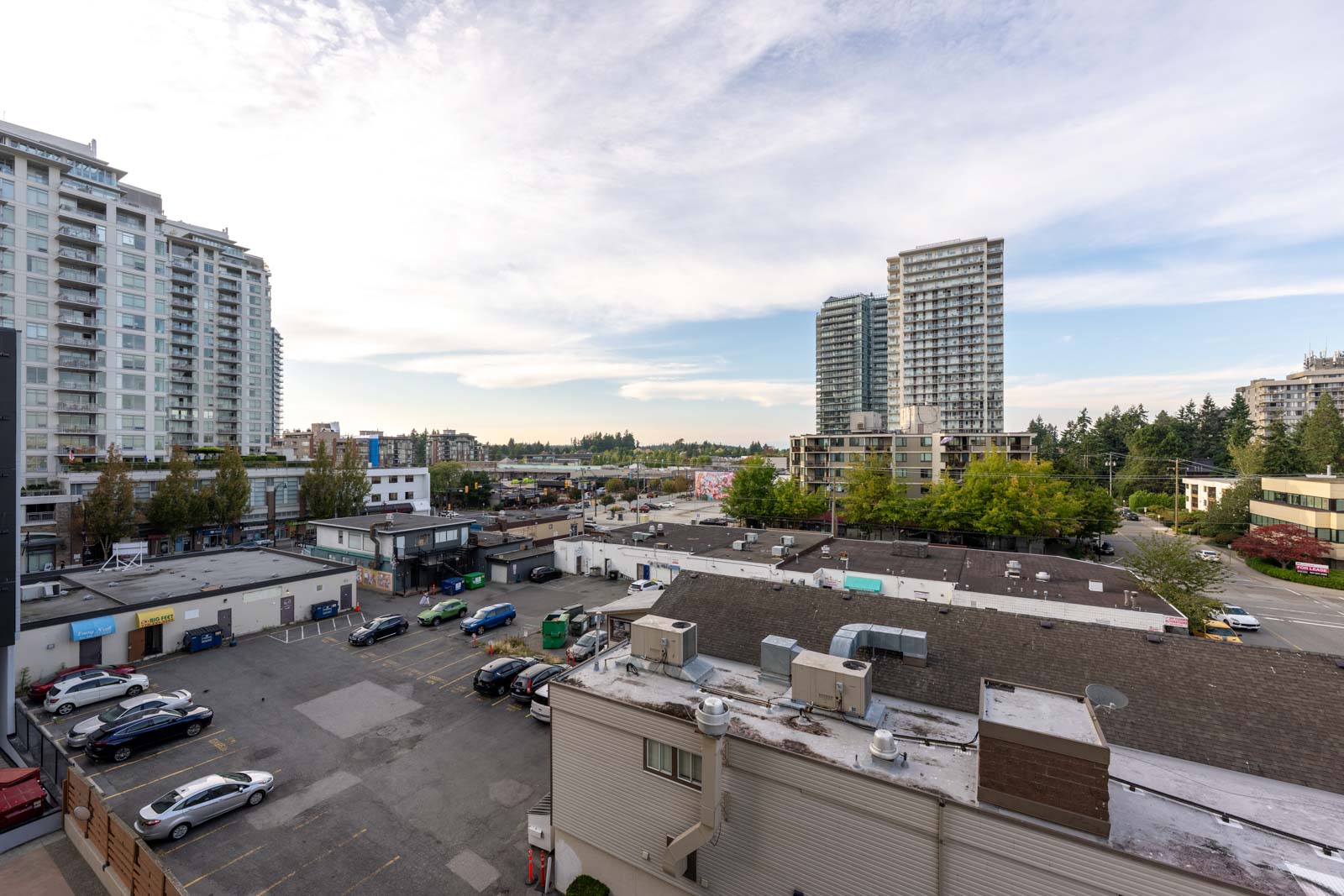View of a cityscape with mid-rise and high-rise buildings, a parking lot with cars, and trees scattered throughout under a partly cloudy sky.