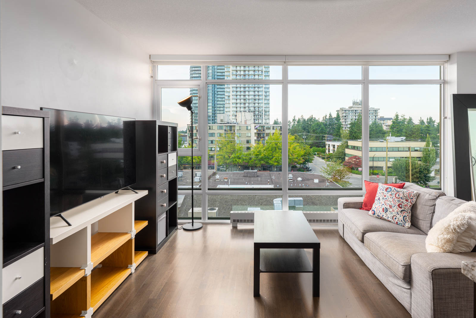 Modern living room with large windows, wooden floor, grey sofa, black coffee table, TV on a white stand, and cityscape view outside.