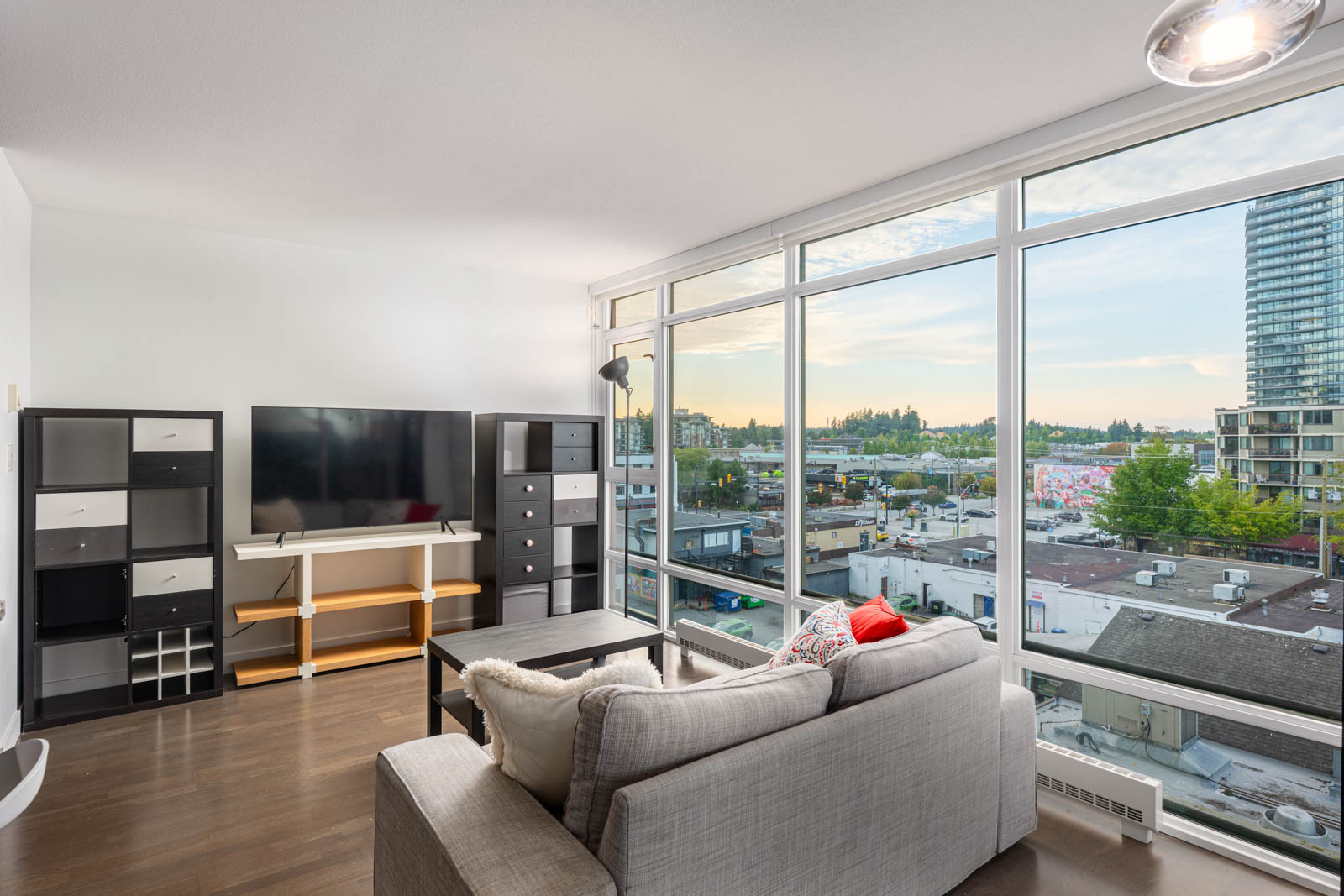Modern living room with gray sofa, TV, shelving units, and large windows overlooking an urban cityscape with buildings and greenery outside.