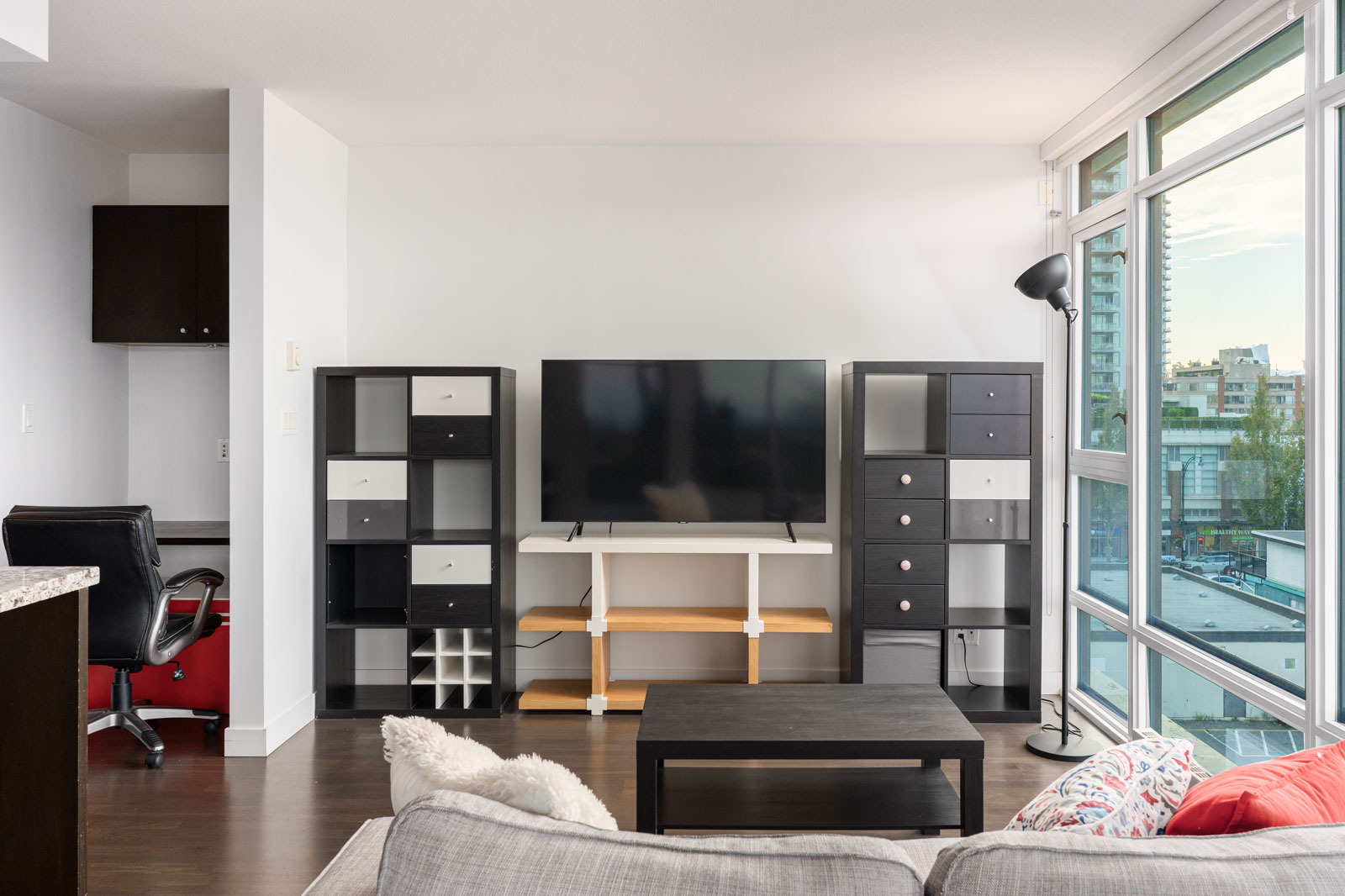 A modern living room with a flat-screen TV on a stand between two black shelving units, a black coffee table, a gray sofa, and large windows overlooking the city.