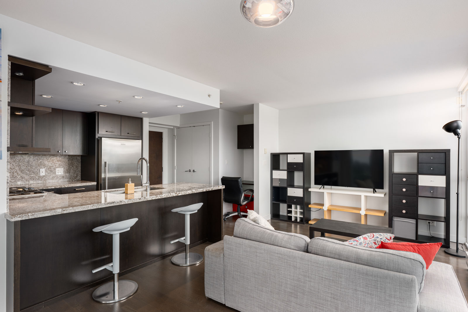 Modern open-plan living room and kitchen with dark cabinets, granite countertop, stools, light gray sofa, TV on a white stand, and bookshelves against a white wall.