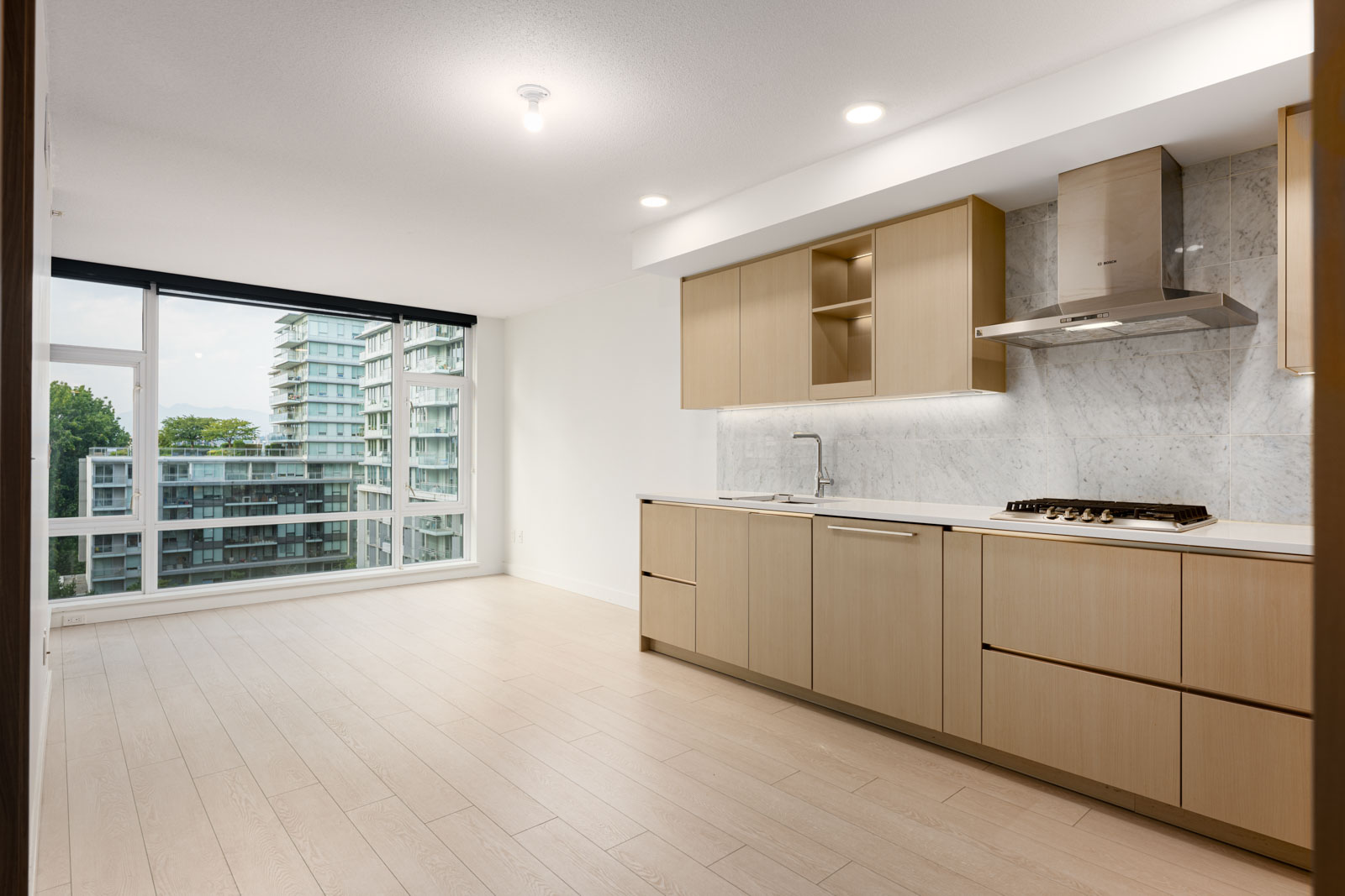 Modern, unfurnished kitchen and dining area with light wood cabinets, built-in appliances, and large window overlooking apartment buildings and greenery.