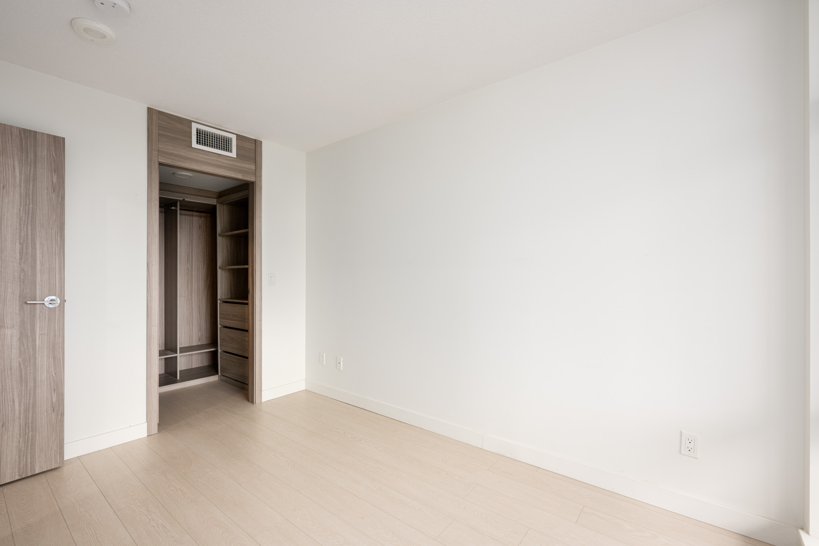 Empty room with light-colored wood flooring, white walls, an open closet with shelves, a closed wooden door, and a power outlet on the right wall.