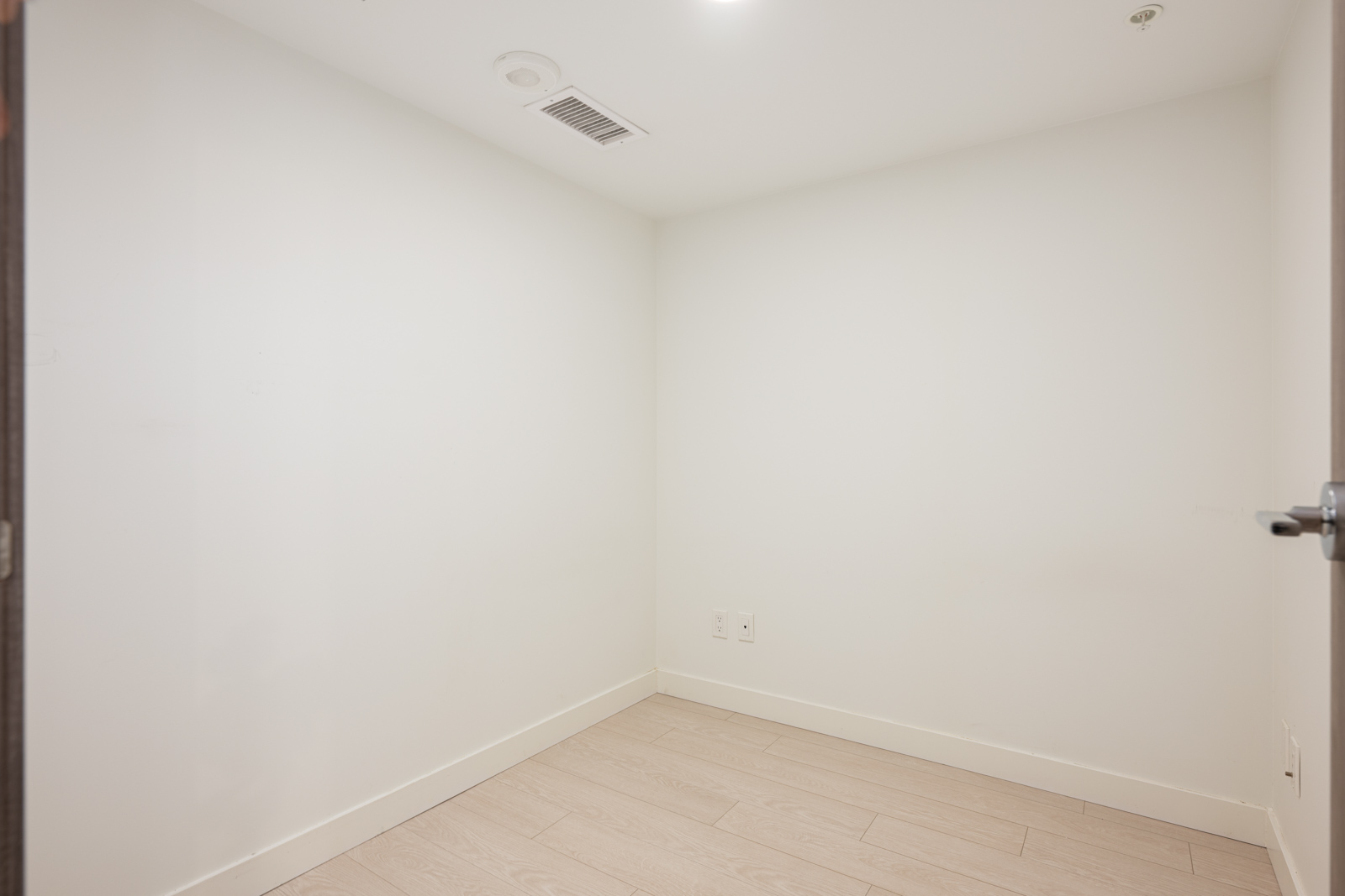Empty, small room with white walls, light wood flooring, a ceiling vent, electrical outlets, and a door slightly open on the right side.