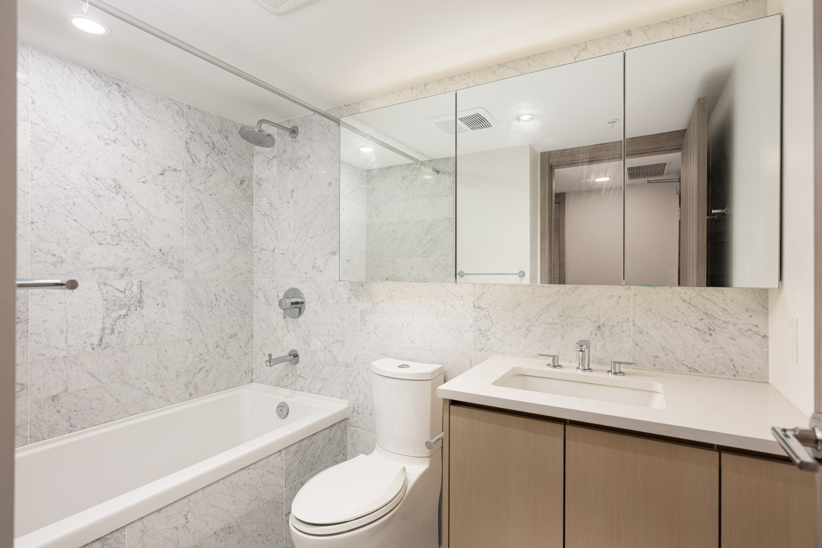 Modern bathroom with a marble-tiled wall, bathtub with shower, toilet, vanity with sink, and mirrored cabinets above the sink.