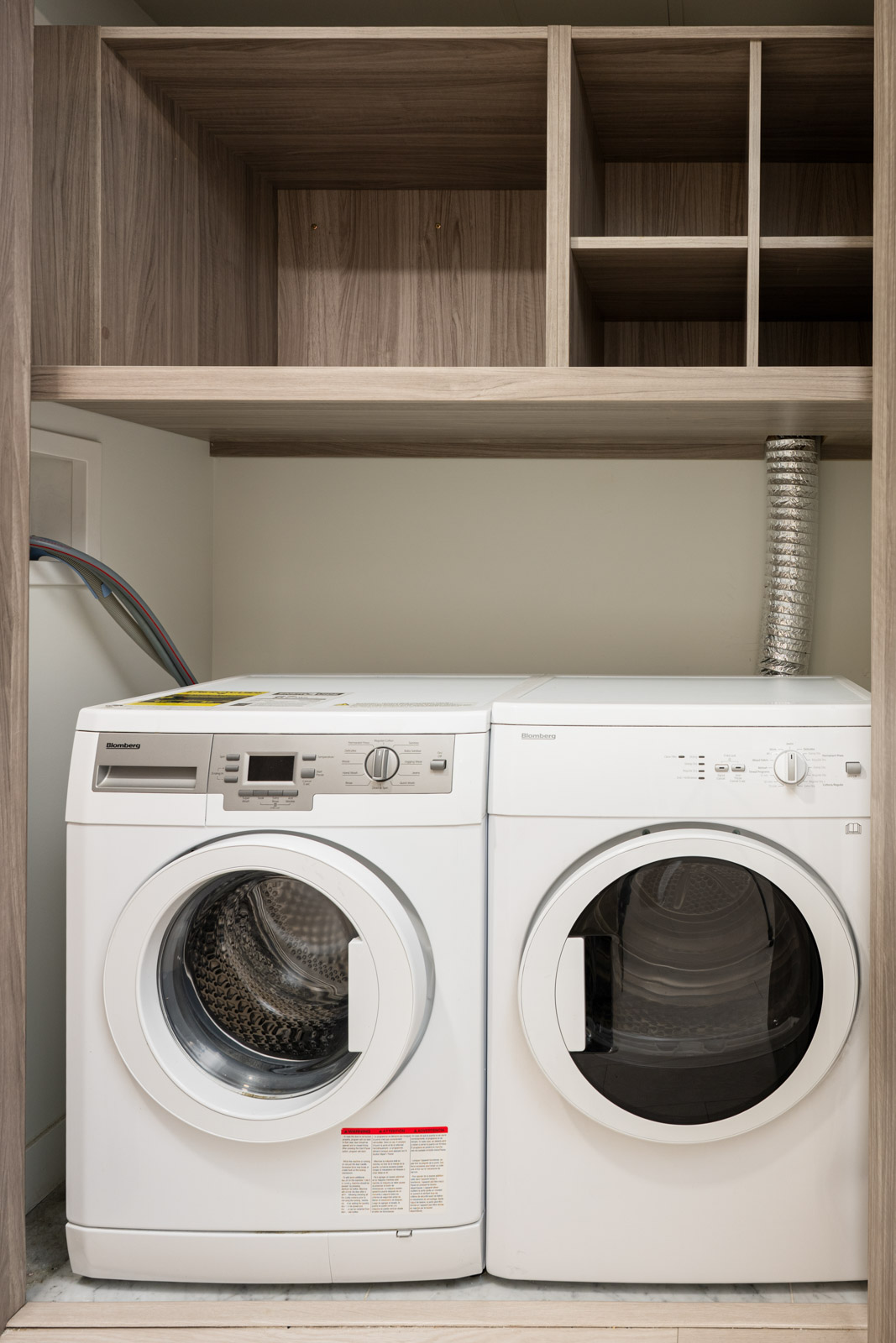 Front-loading washing machine and dryer placed side by side under wooden shelves in a laundry area, with exposed hoses and vent pipe visible.
