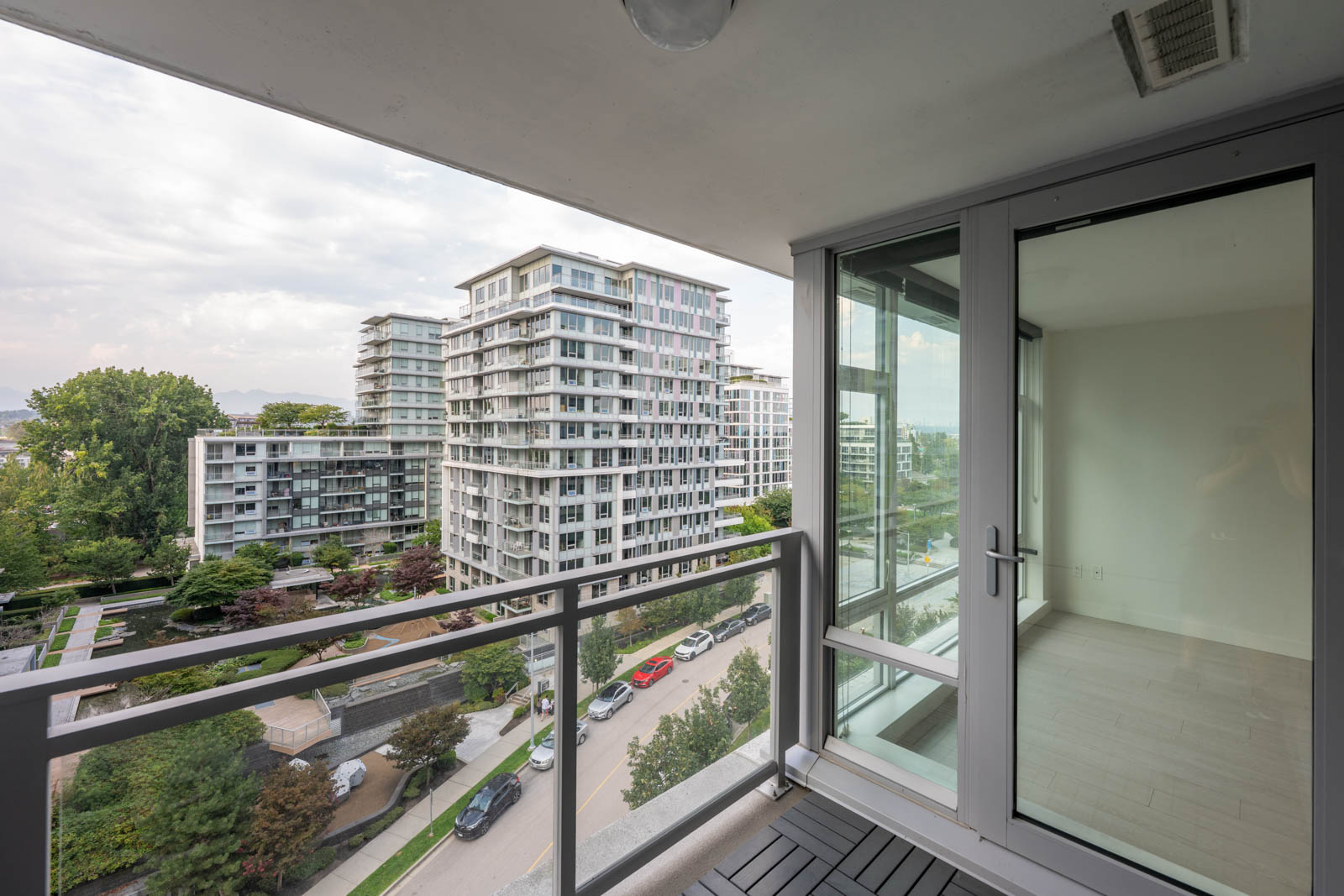 View from a modern apartment balcony overlooking a residential complex with multiple mid-rise buildings, trees, parked cars, and a partially visible empty room through glass doors.