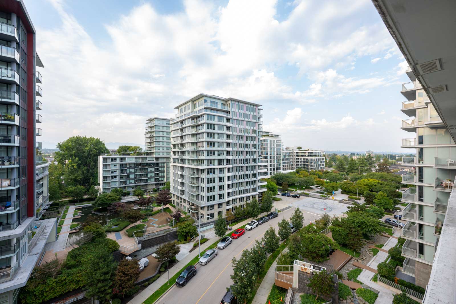 High-rise residential buildings surround a tree-lined street with parked cars and landscaped gardens under a partly cloudy sky.
