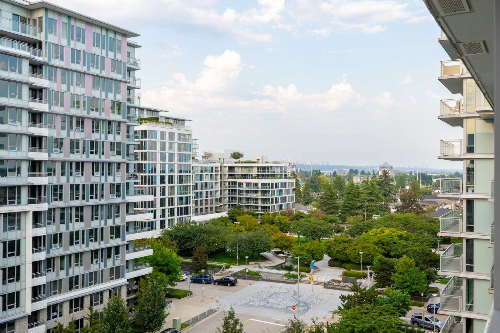 View of modern apartment buildings surrounding a tree-filled courtyard with parked cars and walking paths under a partly cloudy sky.