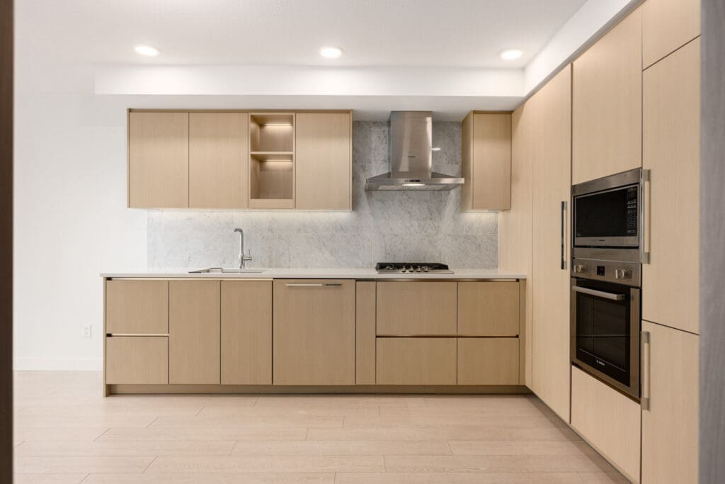 Modern kitchen with light wood cabinets, built-in oven and microwave, gas cooktop with range hood, marble backsplash, and under-cabinet lighting.