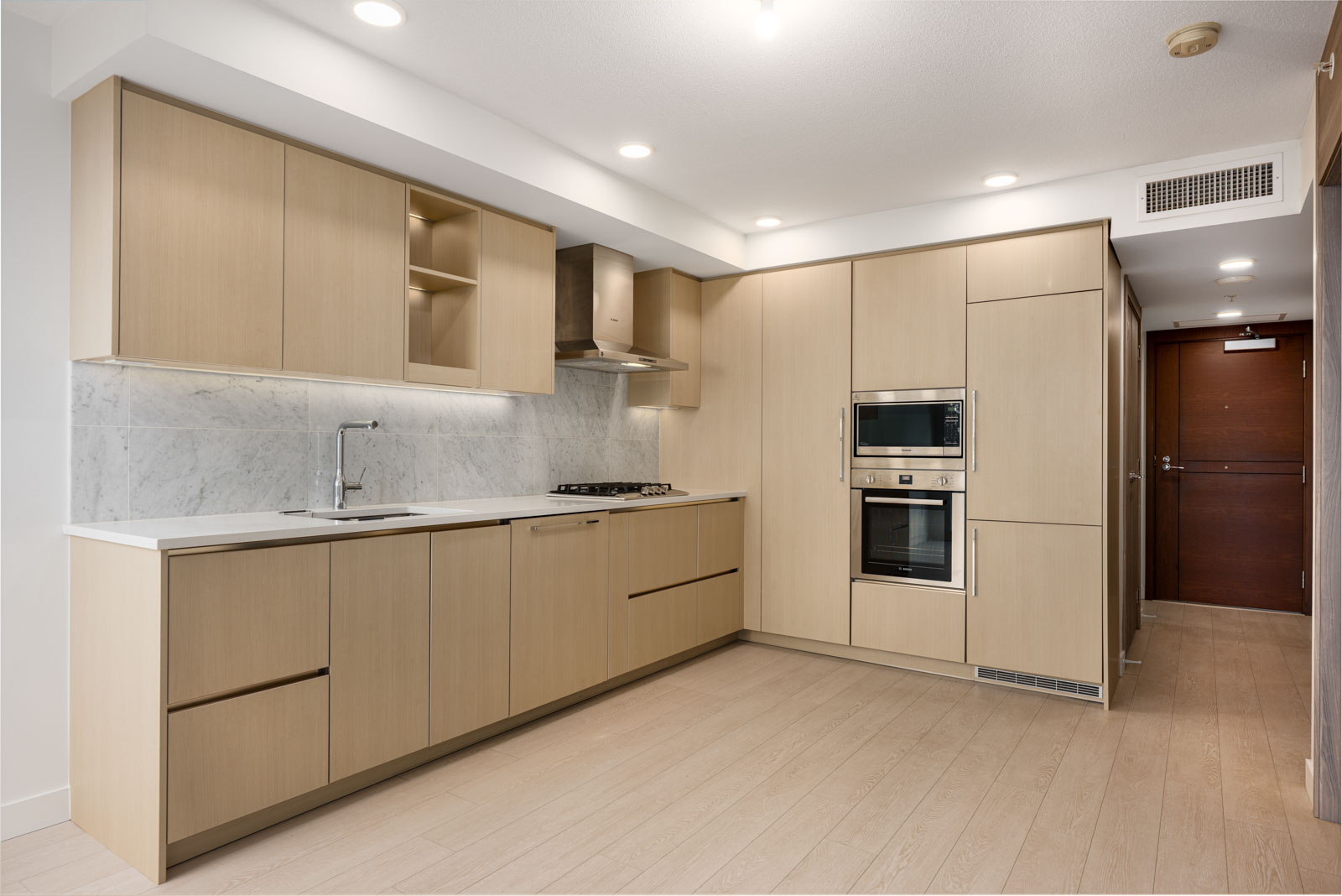 Modern kitchen with light wood cabinets, built-in oven and microwave, marble backsplash, and stainless steel appliances. The room has light wood flooring and recessed lighting.