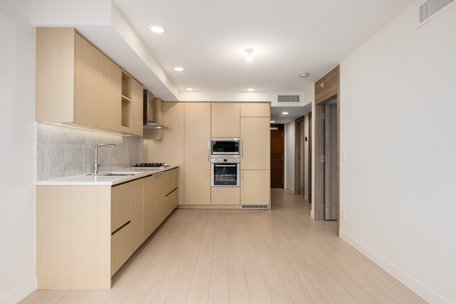 Modern kitchen with light wood cabinets, built-in appliances, marble backsplash, and light wood flooring, adjacent to a hallway leading to other rooms.
