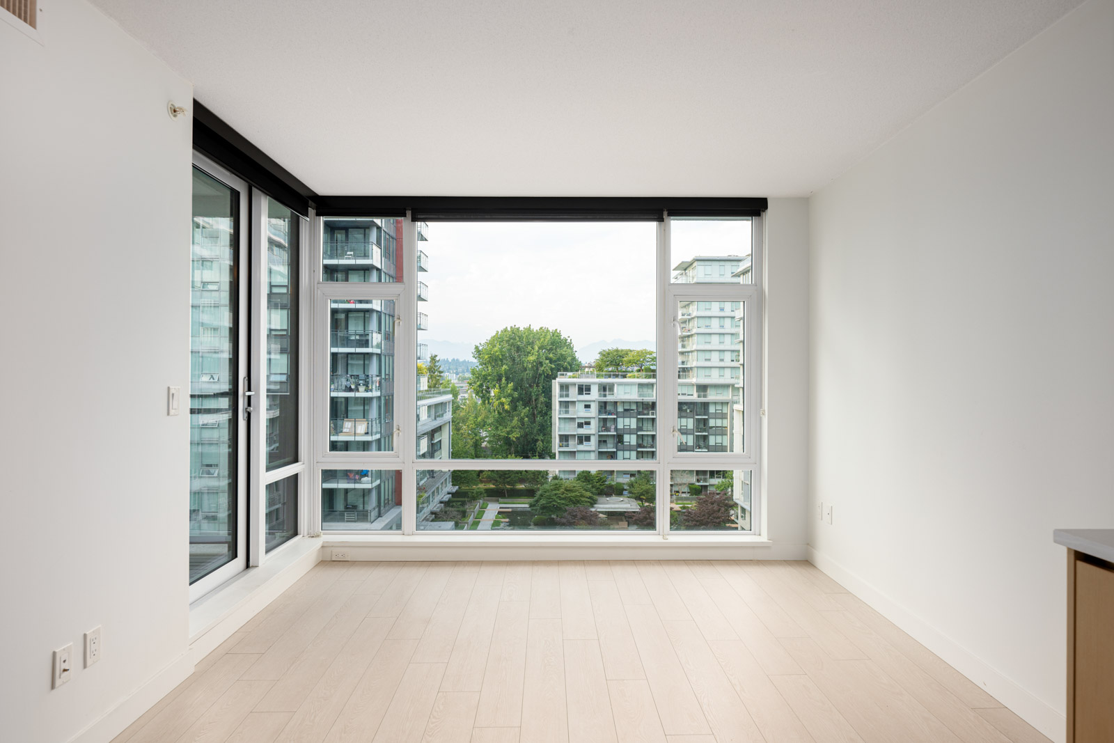 Unfurnished room with large windows, light wood floors, white walls, and a view of modern apartment buildings and greenery outside.