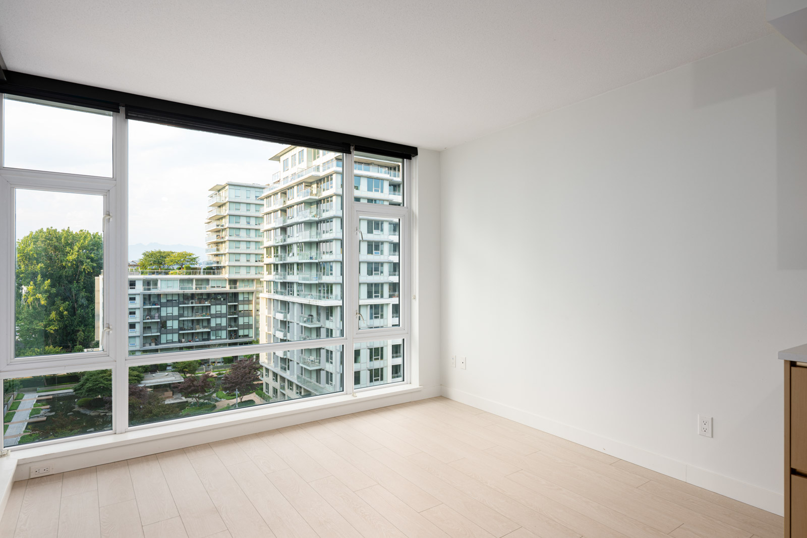 Empty modern room with light wood flooring, white walls, and large floor-to-ceiling windows showing a view of residential apartment buildings and greenery outside.
