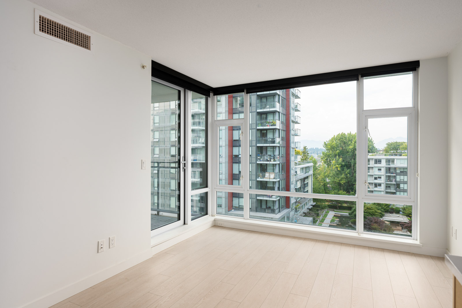 Unfurnished corner apartment room with large floor-to-ceiling windows, light wood flooring, and views of modern apartment buildings and greenery outside.