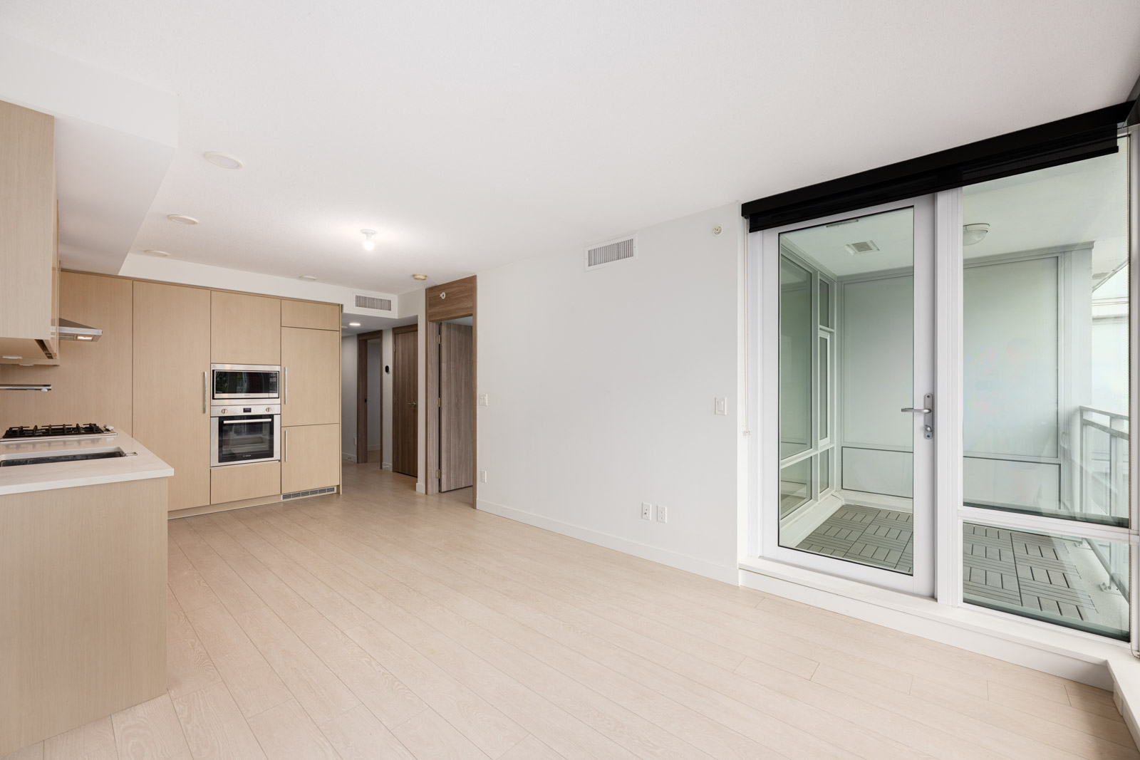 Modern, unfurnished apartment interior with light wood floors, built-in kitchen appliances, and large sliding glass door leading to an enclosed balcony or sunroom.