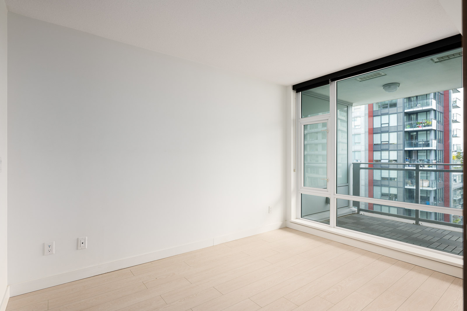 Unfurnished room with light wood flooring, white walls, electrical outlets, and a large window leading to a balcony with views of nearby apartment buildings.