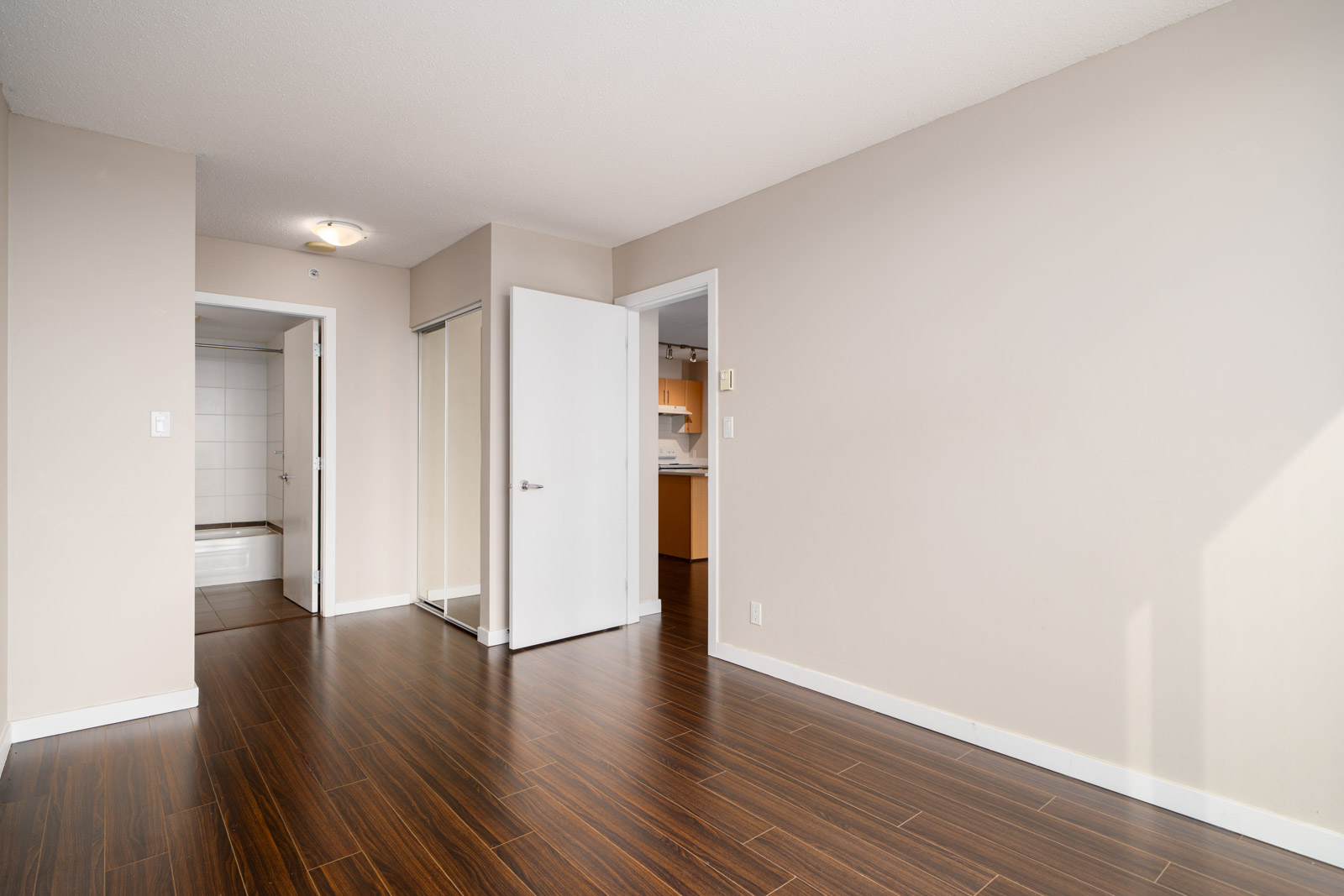 Empty room with beige walls, dark wood flooring, an open closet with mirrored doors, and view into a kitchen and bathroom area through open doors.