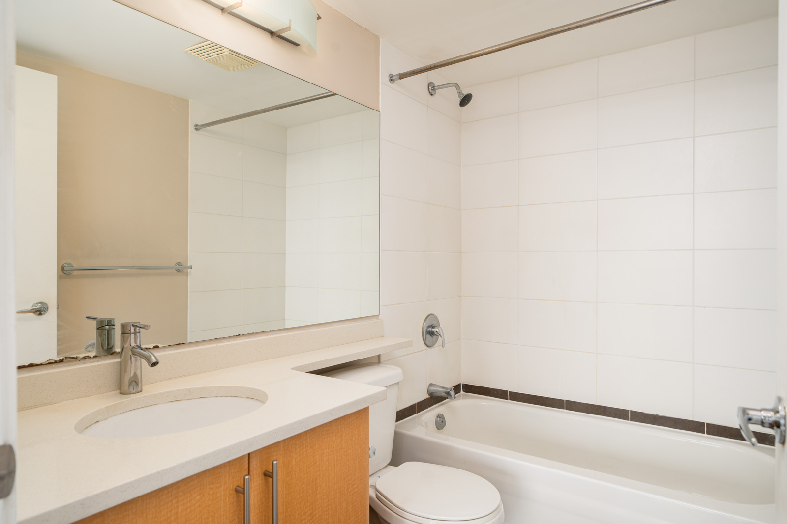 Bathroom with a large mirror, sink with countertop, wooden vanity, toilet, and a bathtub with a shower curtain rod and white tiled walls.