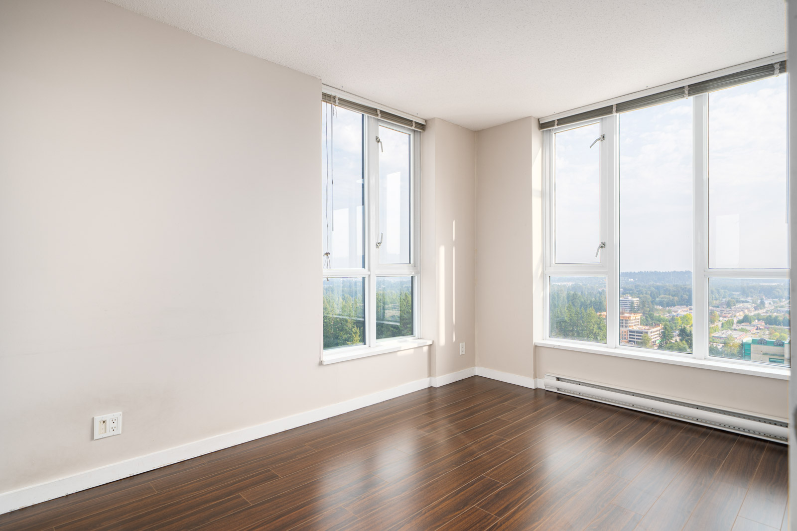 Empty room with light beige walls, large windows letting in natural light, and dark wood flooring. Cityscape and greenery are visible outside the windows.
