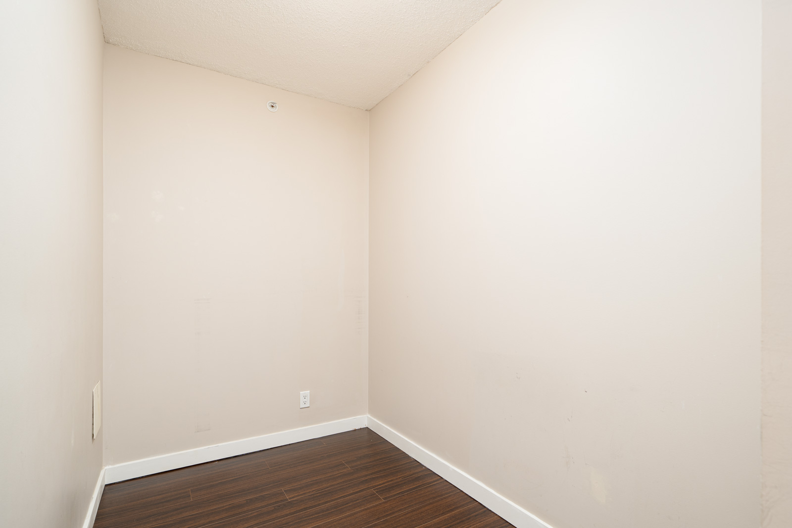 Small, empty room with beige walls, white baseboards, and dark wood flooring. No furniture or decor is present.