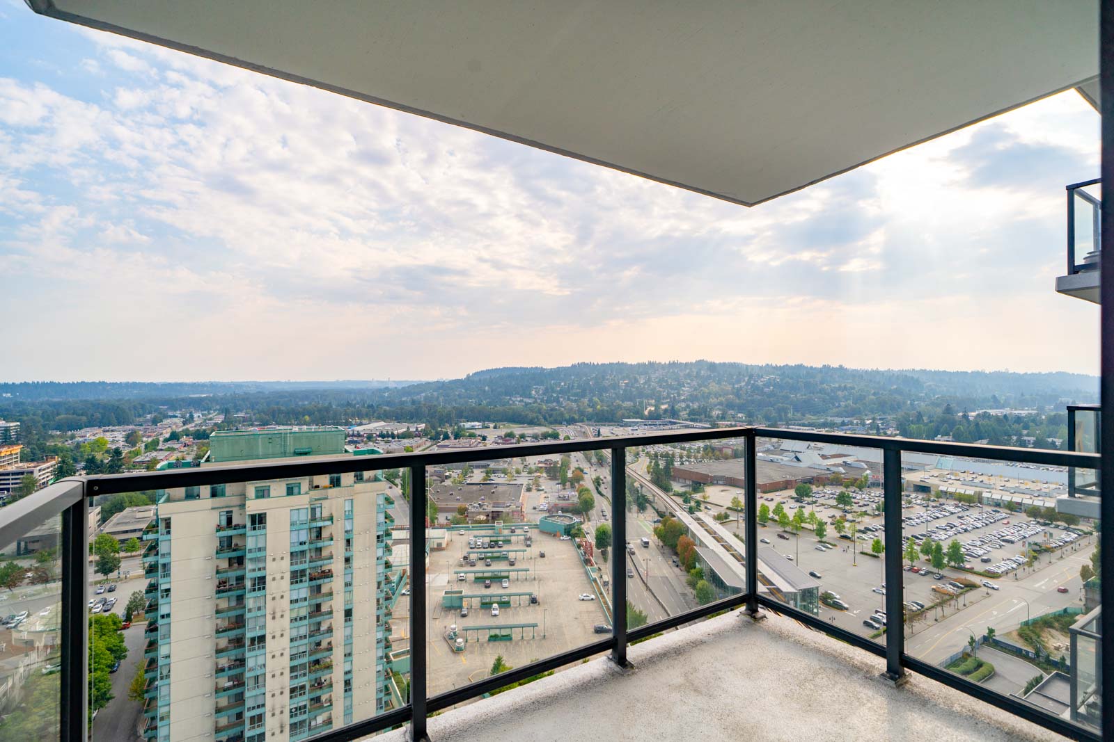View from a high-rise balcony overlooking a cityscape with buildings, roads, and a distant tree-covered hill under a partly cloudy sky.