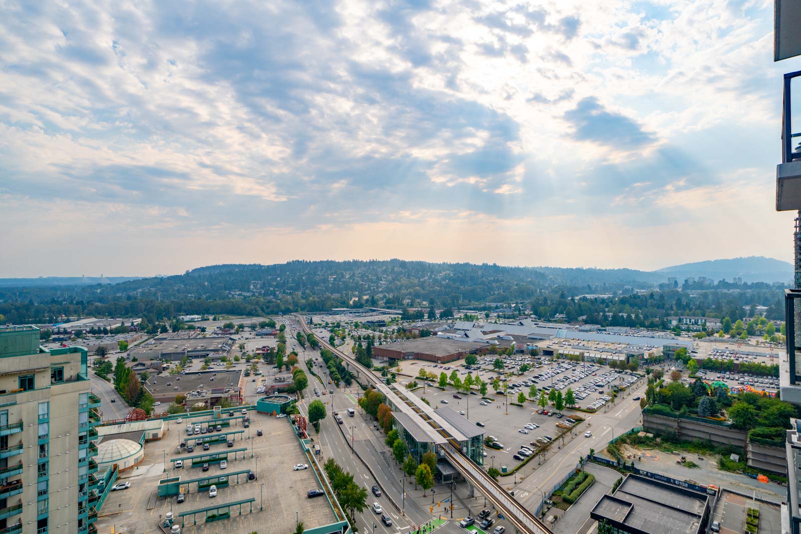 Aerial view of an urban area with roads, buildings, parking lots, and a railway station, backed by tree-covered hills under a partly cloudy sky.