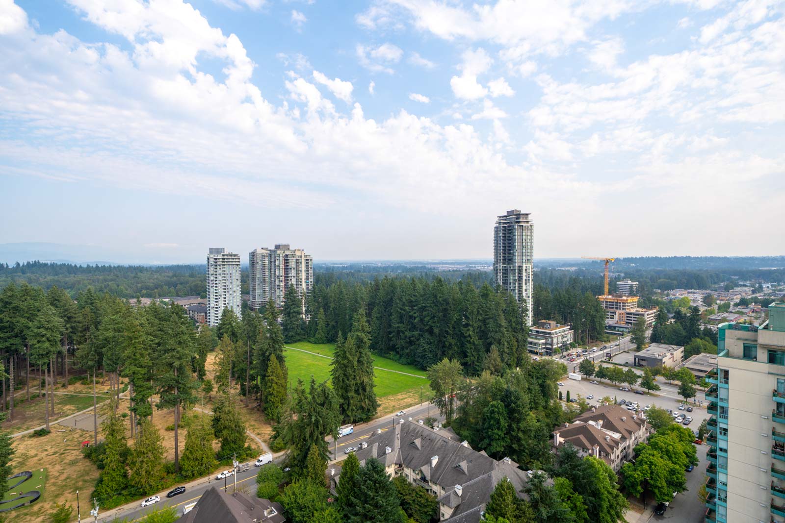 Aerial view of a cityscape with high-rise buildings, residential houses, a sports field, and dense green trees under a partly cloudy sky.