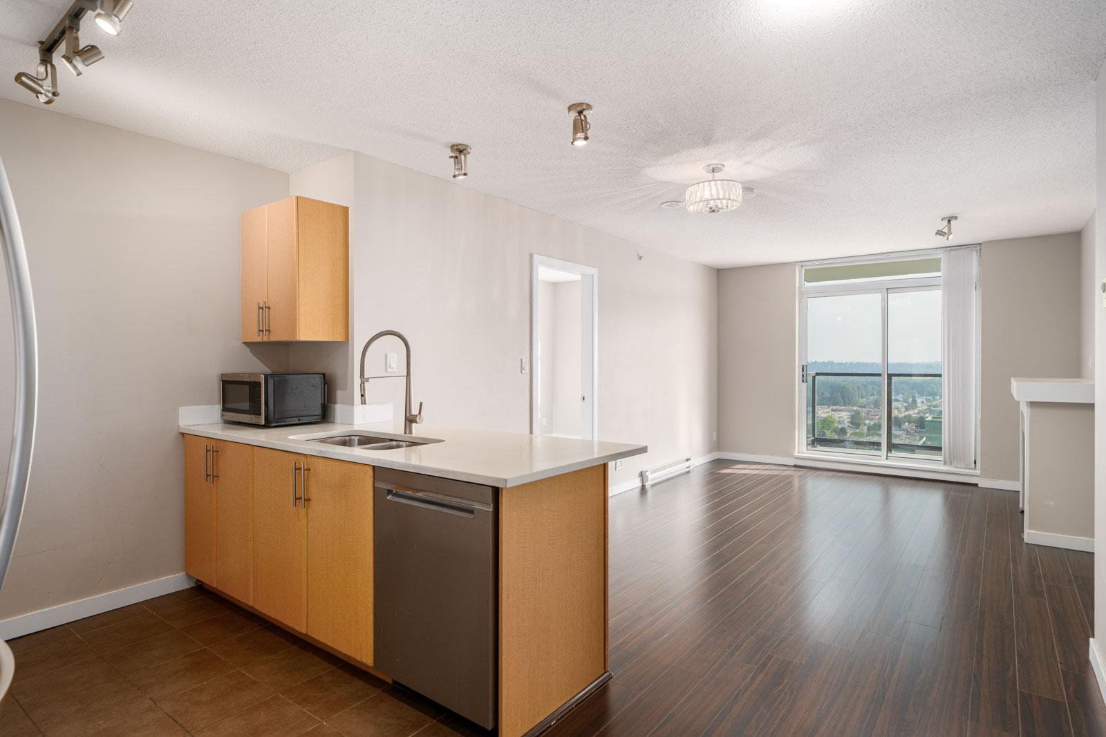 Modern apartment kitchen with light wood cabinets, stainless steel appliances, and open living area featuring large window and dark wood flooring.