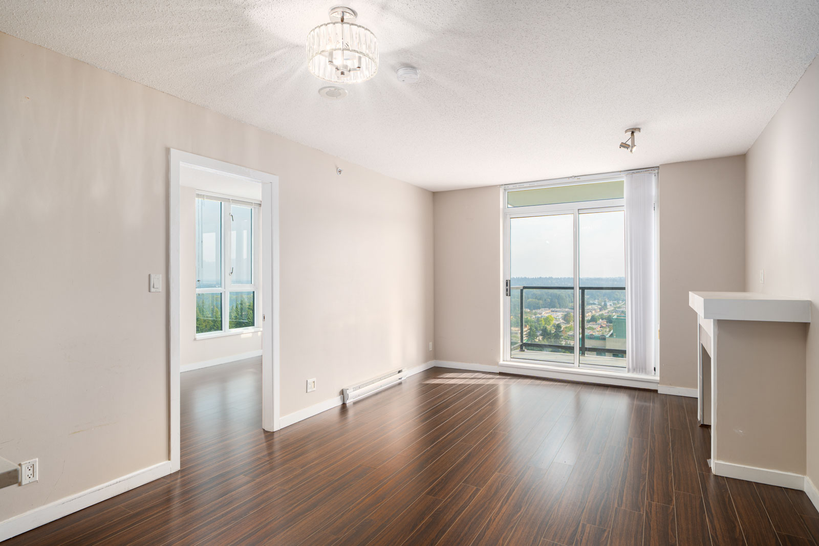 Unfurnished room with wood flooring, beige walls, a ceiling light, large window with balcony and view, small fireplace, and an adjoining room through a doorway.