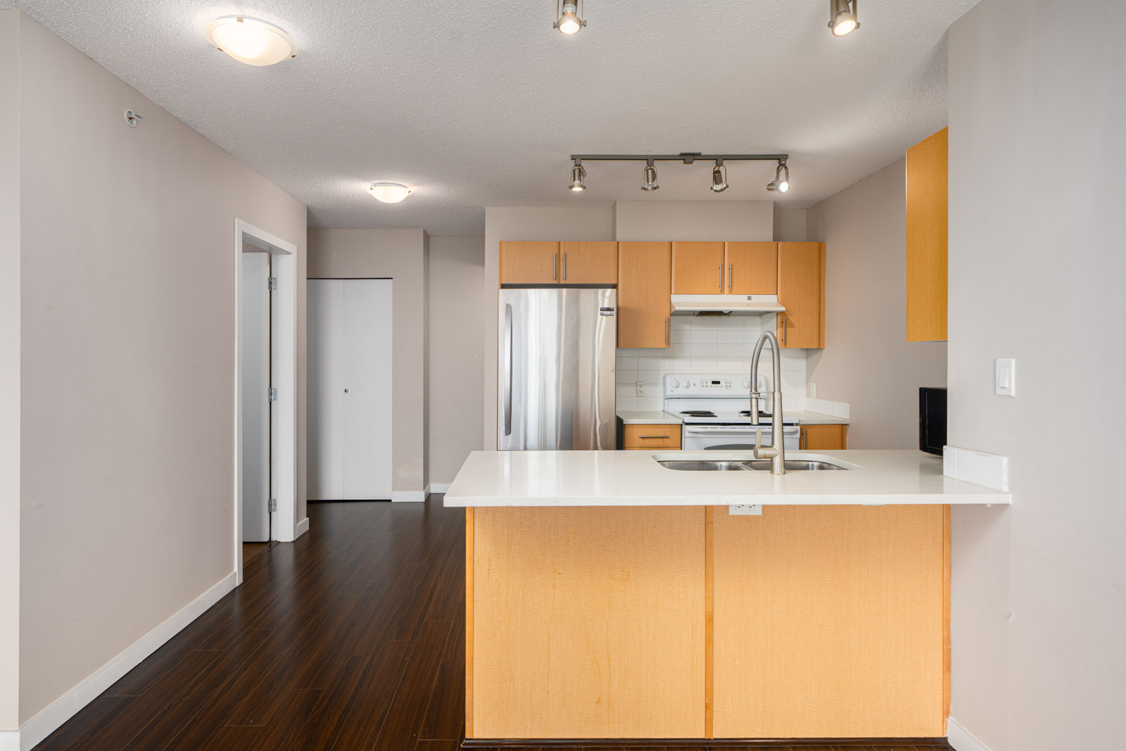 Modern kitchen with light wood cabinets, white countertops, stainless steel refrigerator, oven, and sink, featuring track lighting and dark wood flooring.