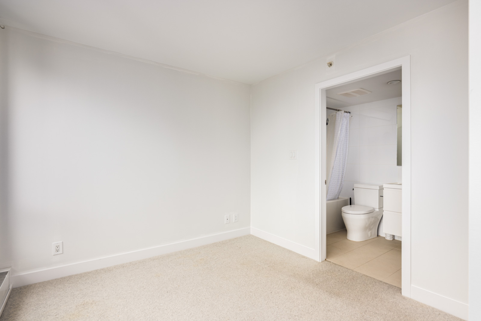 Unfurnished room with beige carpet and white walls, adjacent to a bathroom with a toilet, sink, and white shower curtain visible through an open doorway.