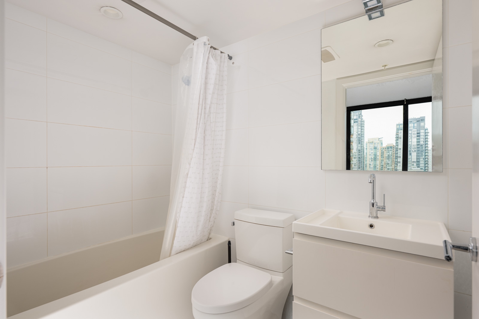 Modern bathroom with a white bathtub and shower curtain, a toilet, a sink with a cabinet, and a large mirror reflecting a window view of city buildings.