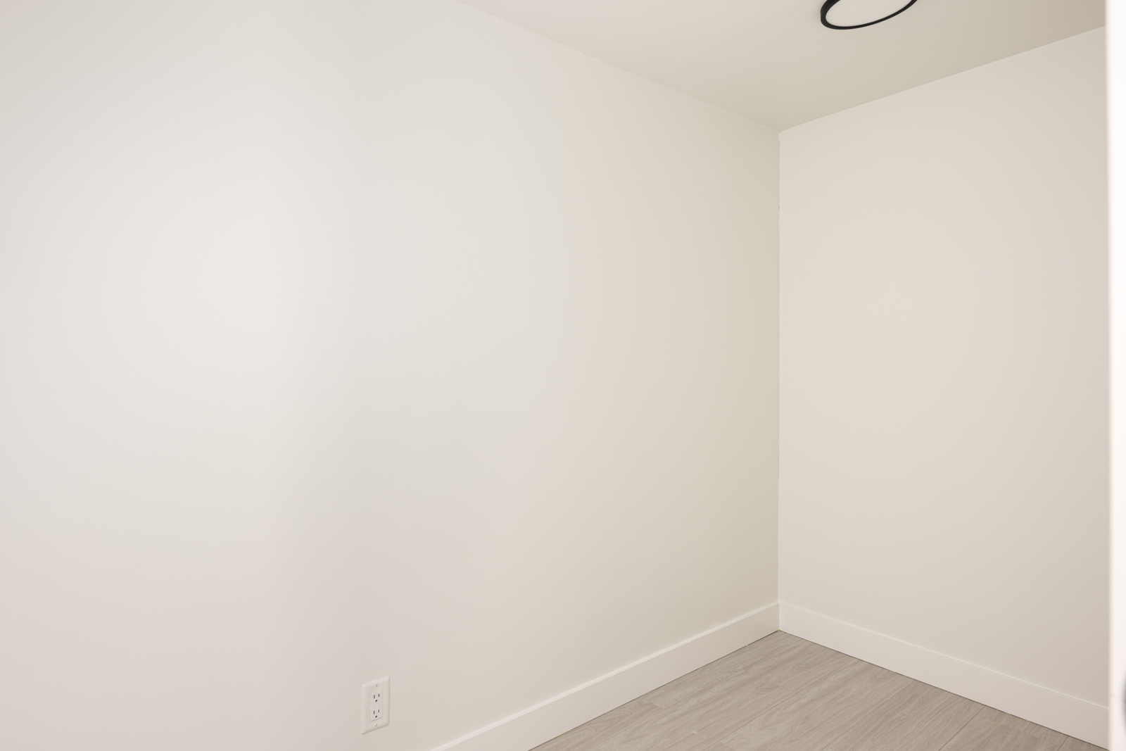 Empty white room with plain walls, light-colored flooring, baseboard trim, electrical outlet, and a ceiling fixture partially visible.