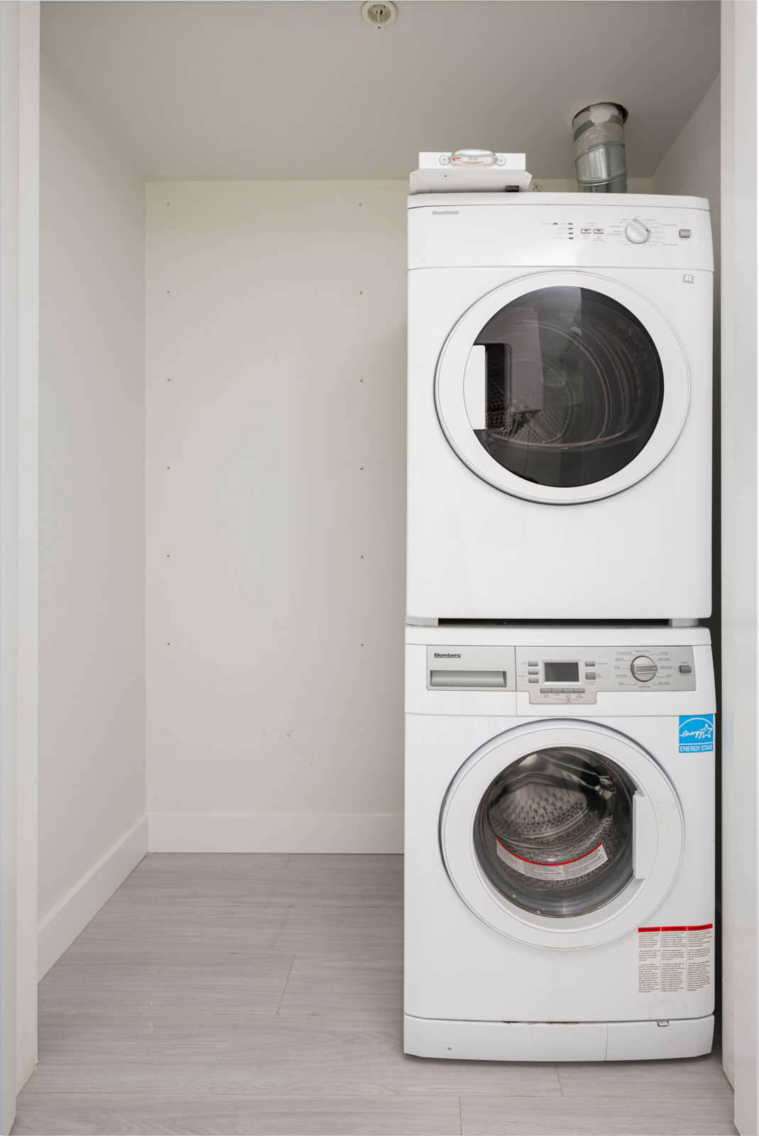 A stacked white washer and dryer set is positioned in a small laundry nook with light-colored flooring and plain white walls.