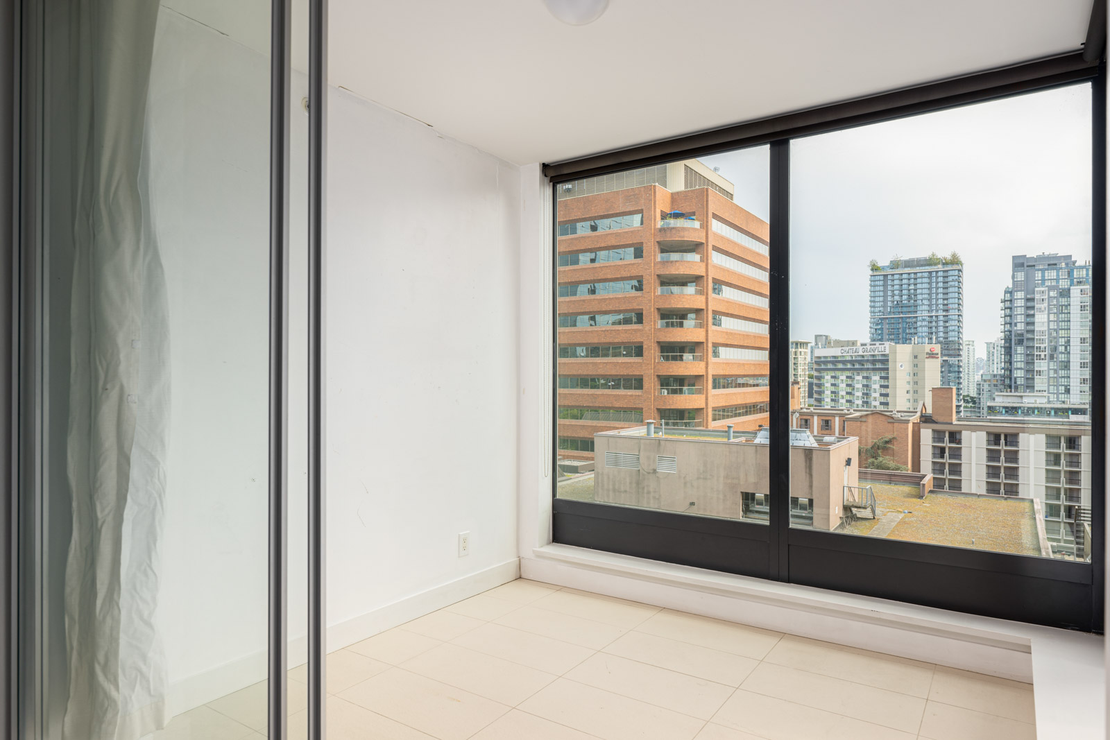 Small empty room with large floor-to-ceiling windows overlooking city buildings, including a tall brown office building and modern high-rises.