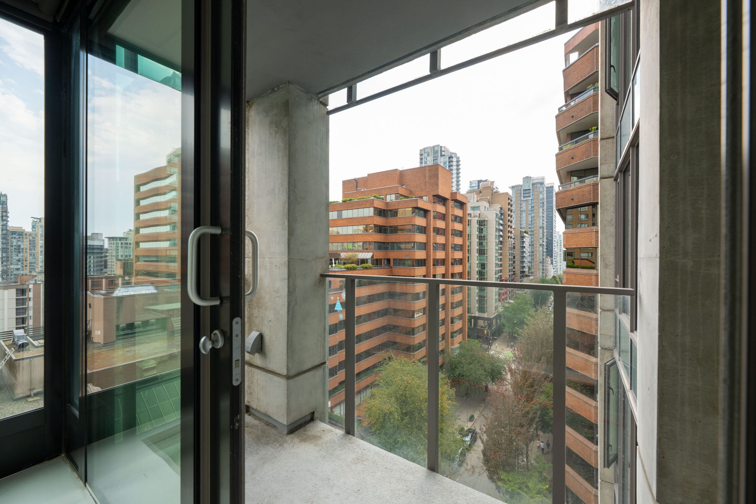 A small balcony with glass railings overlooks a cityscape of modern high-rise buildings and a tree-lined street below.