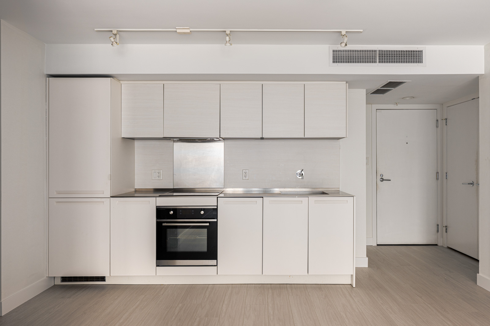 Minimalist kitchen with white cabinets, electric stove, oven, and stainless steel backsplash; light wood flooring and a white entry door are also visible.