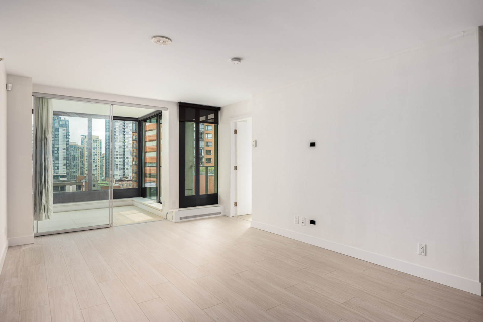 Unfurnished modern apartment room with light wood flooring, white walls, a large window door leading to a balcony, and a view of city buildings.