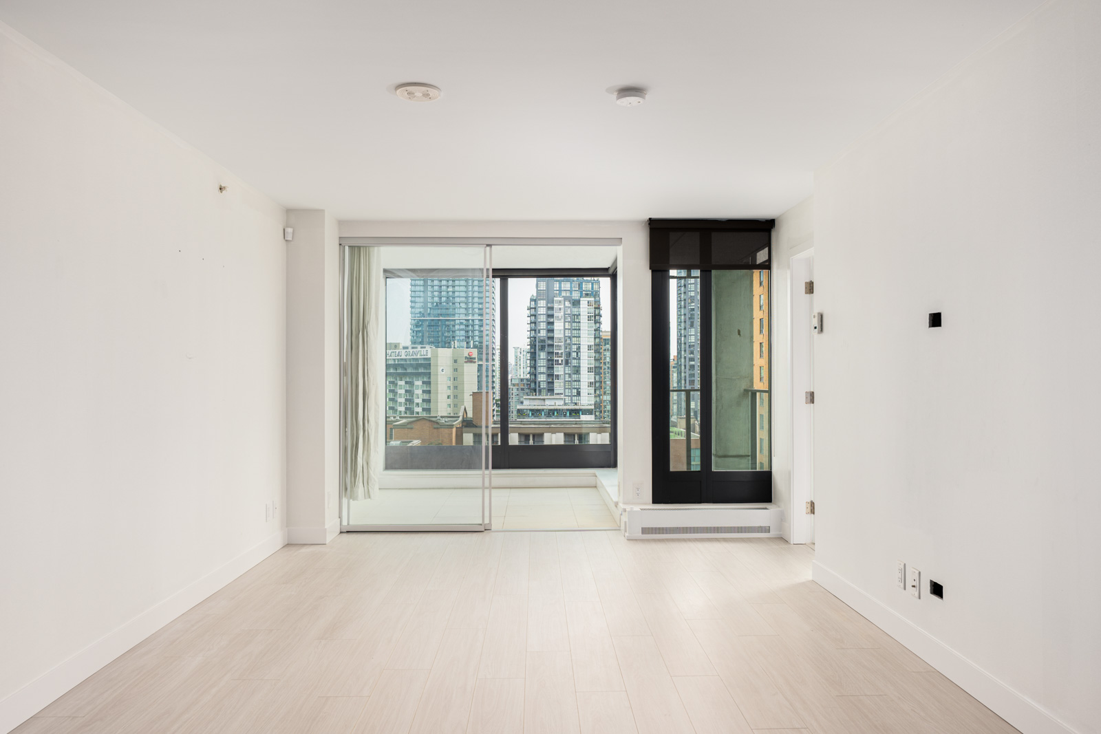 Empty modern apartment room with light-colored flooring, white walls, and large glass doors leading to a balcony with a cityscape view.
