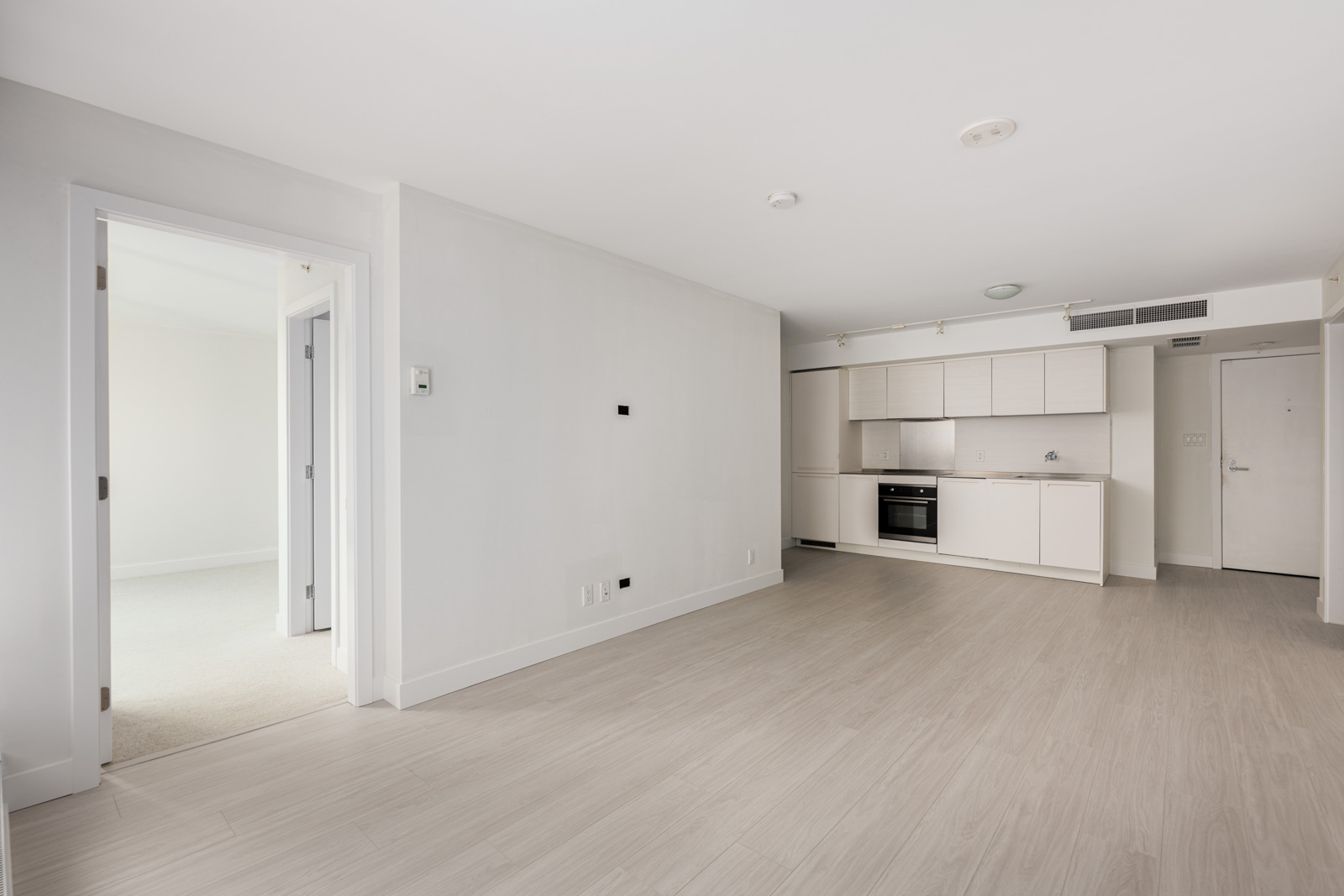 Unfurnished modern apartment with light wood flooring, white walls, an open-plan kitchen, and a separate bedroom visible through an open doorway.