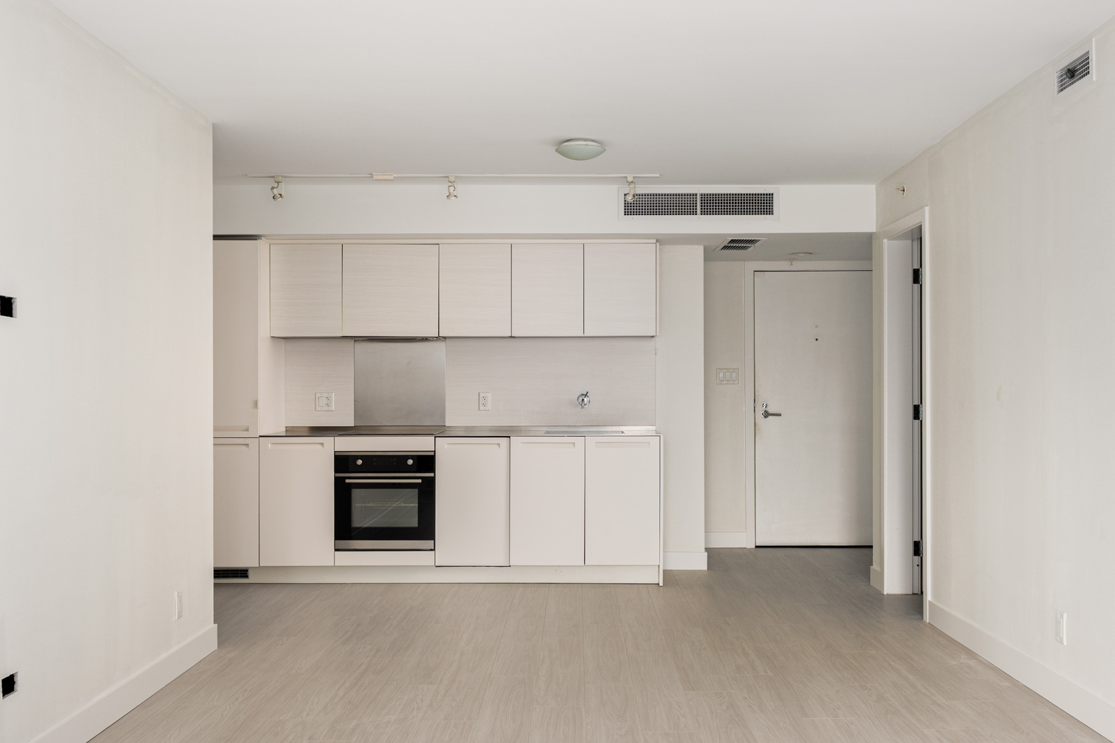 Modern unfurnished apartment with a small kitchen, light wood floors, white cabinets, and a closed entry door visible in the background.