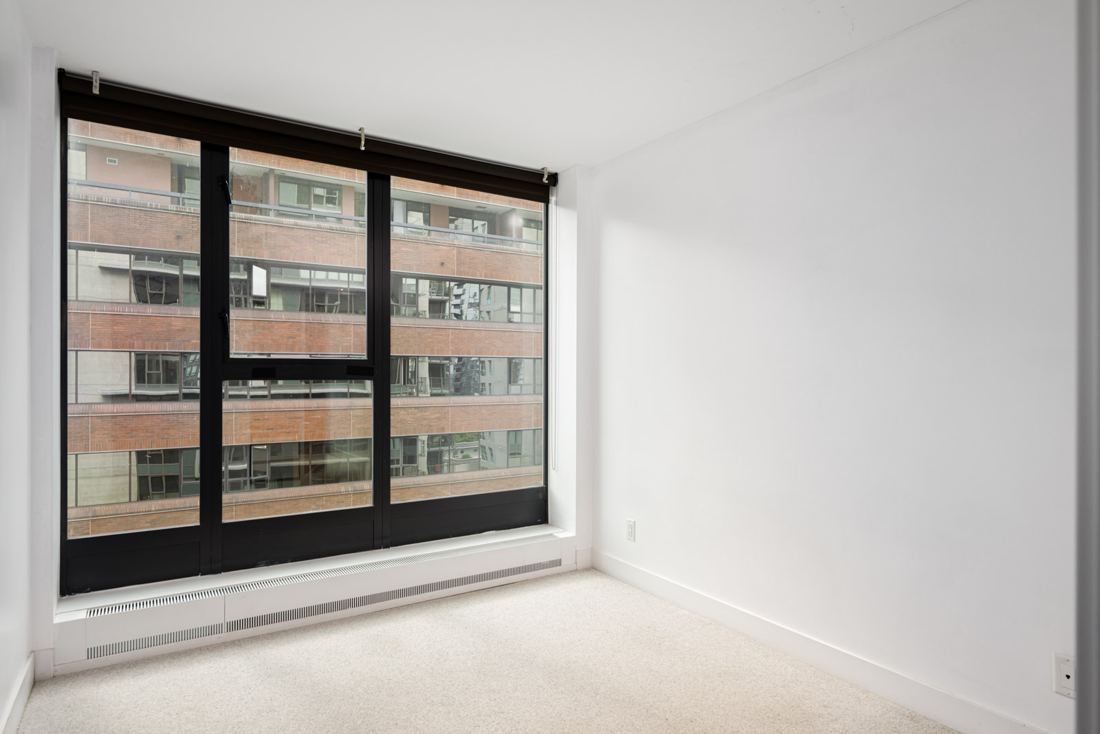Empty room with white walls, beige carpet, and a large floor-to-ceiling window overlooking a neighboring brick building.