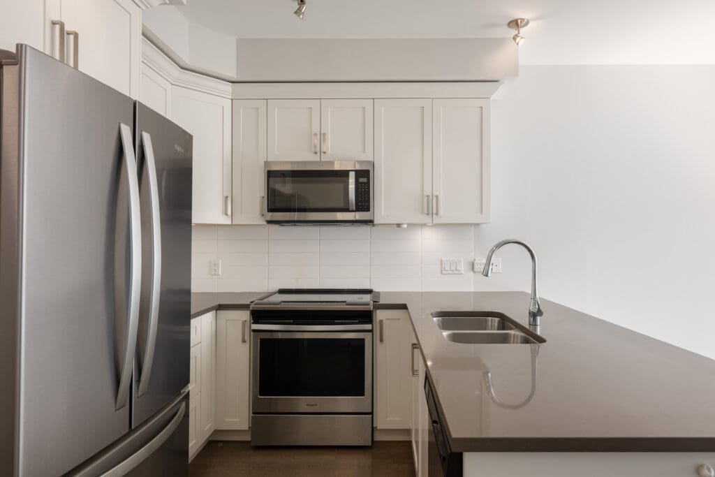 Modern kitchen with stainless steel appliances, white cabinets, a built-in microwave, electric stove, double-door fridge, and a gray countertop with a sink.