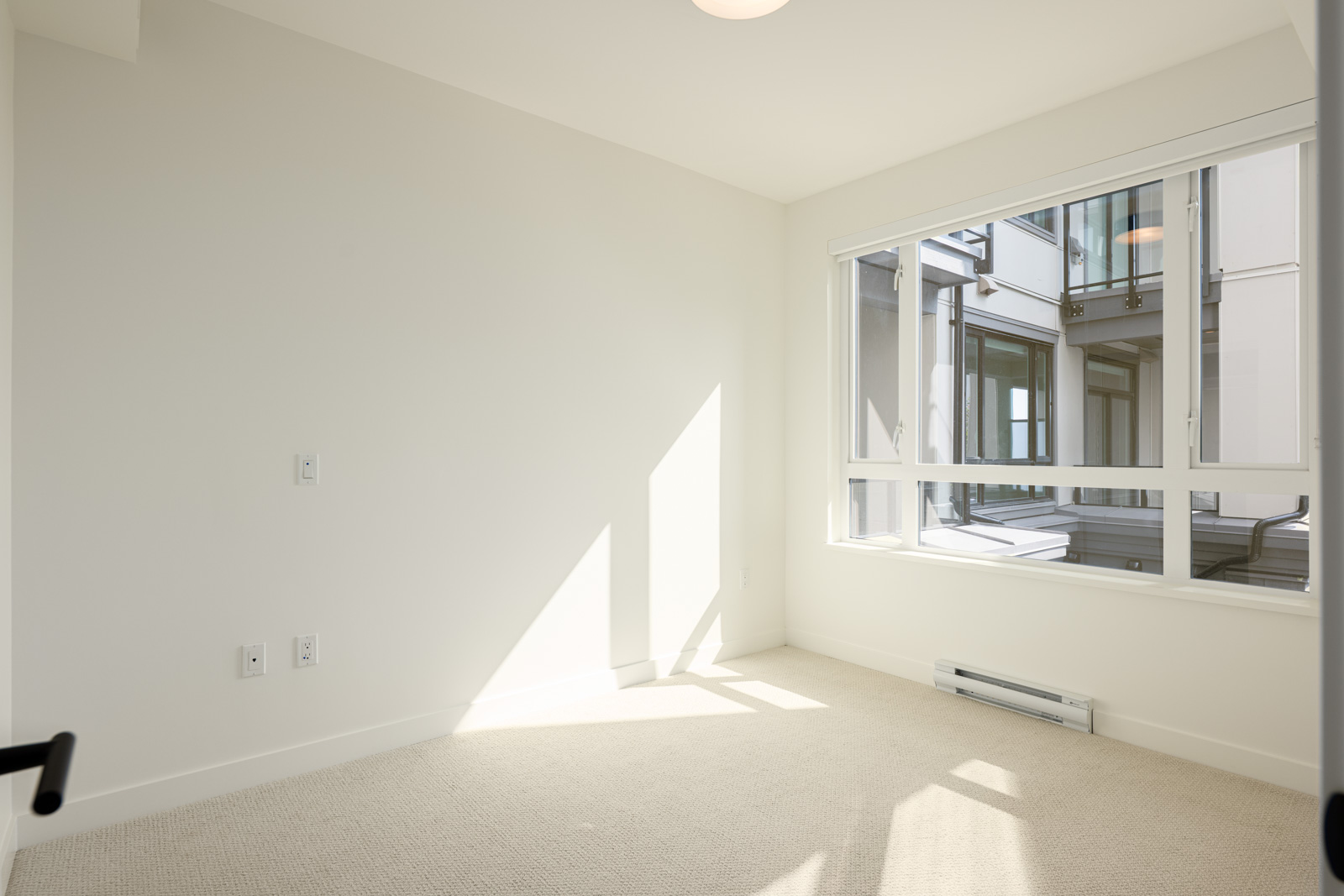 Empty room with beige carpet, white walls, electrical outlets, and a large window letting in natural light, showing an exterior building view.