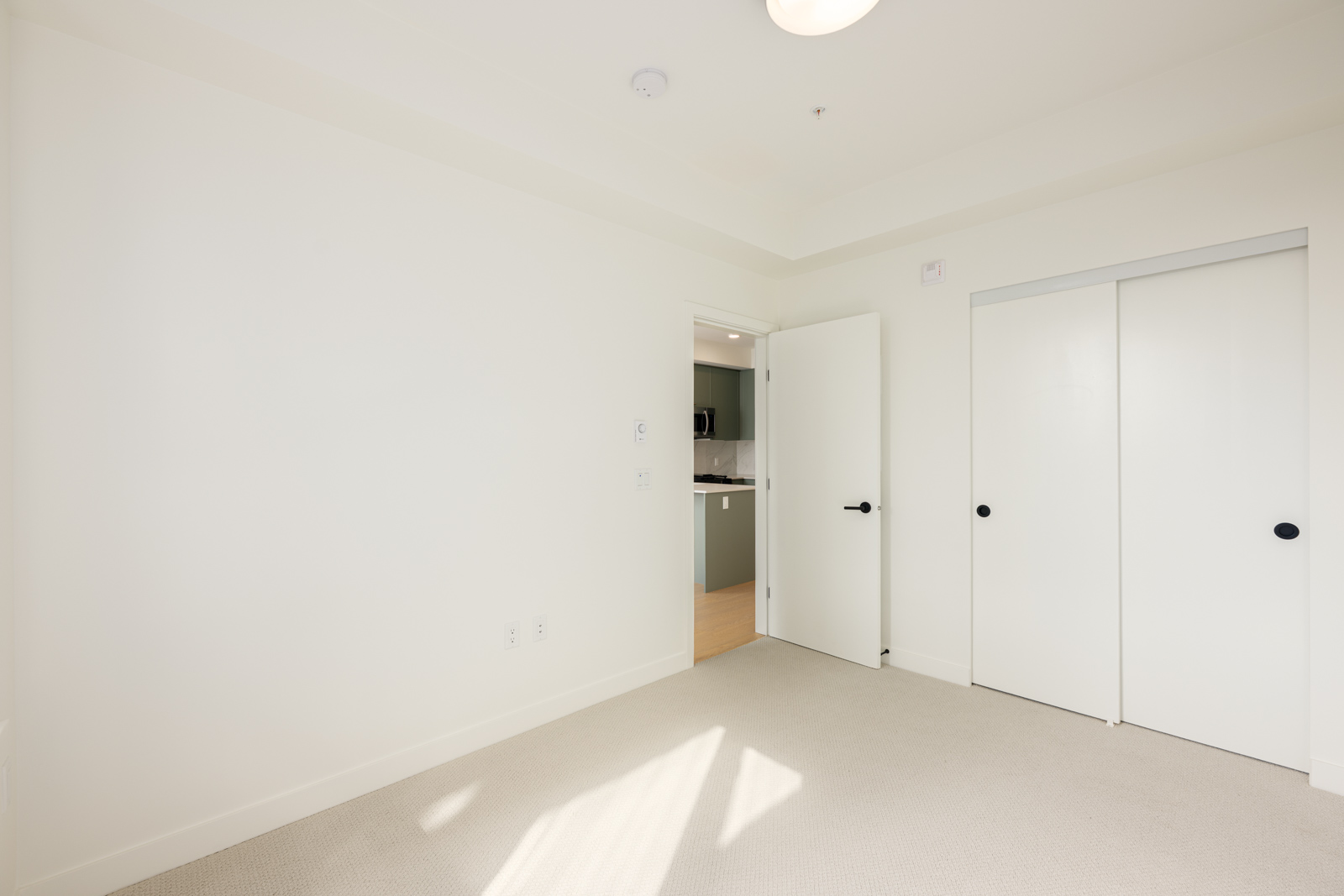 Empty room with white walls, beige carpet, a double sliding closet door, and an open door leading to a kitchen area with visible countertops and cabinets.