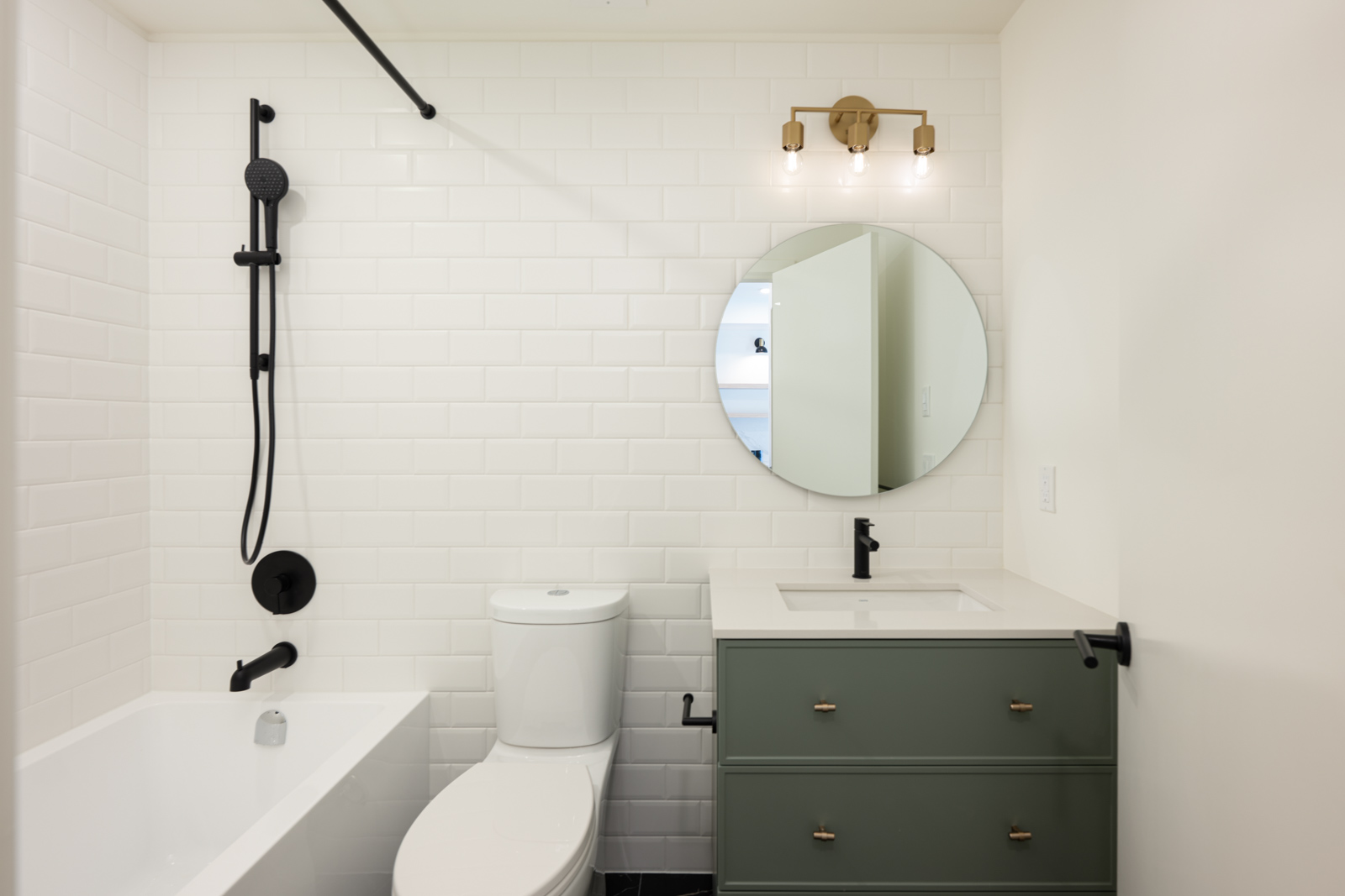 Modern bathroom with white subway tile walls, a round mirror, green vanity with sink, black fixtures, and a toilet beside a bathtub with a black showerhead.