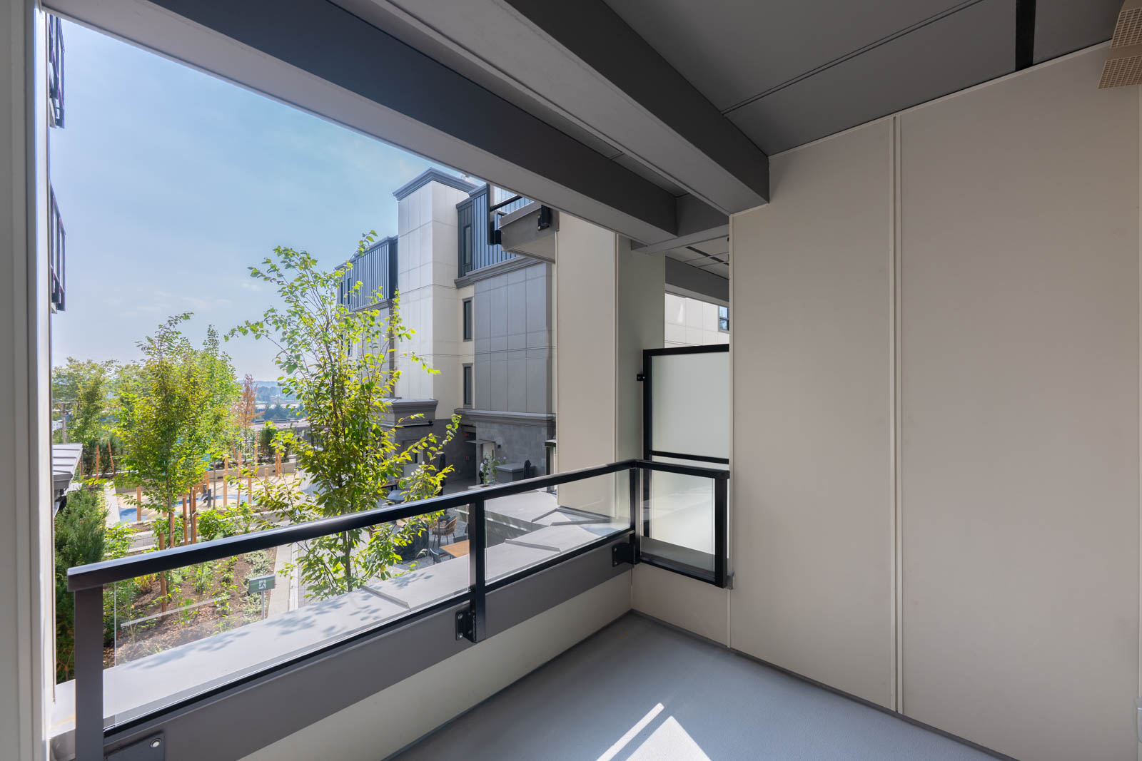 A modern apartment balcony with glass and metal railings overlooks a landscaped area and neighboring buildings under a clear sky.