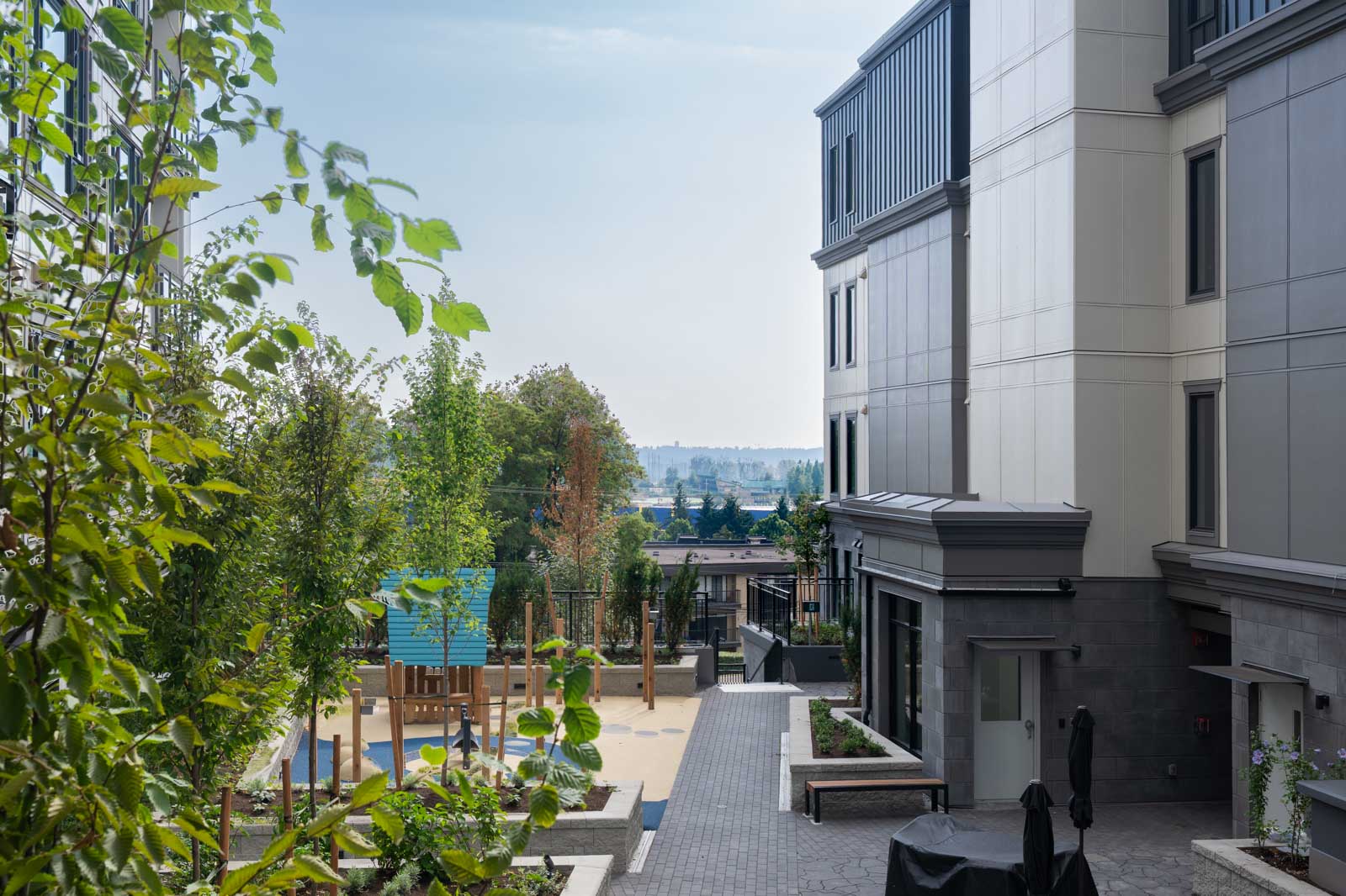 Modern apartment building courtyard with landscaping, seating areas, and a playground, overlooking a distant cityscape under a clear sky.